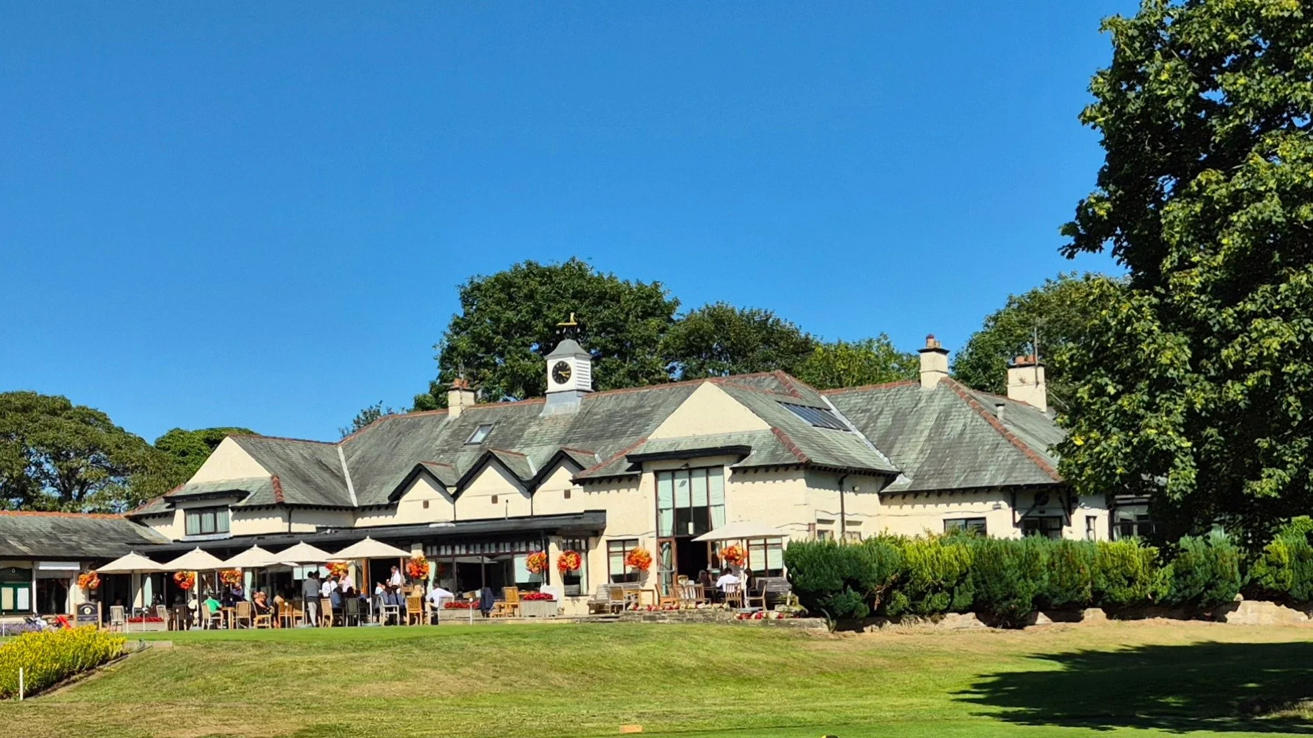 A large cafe or restaurant building with a scenic outdoor patio, white patio umbrellas, and decorated with orange and yellow flowers, set against a backdrop of lush green trees and a bright blue sky.