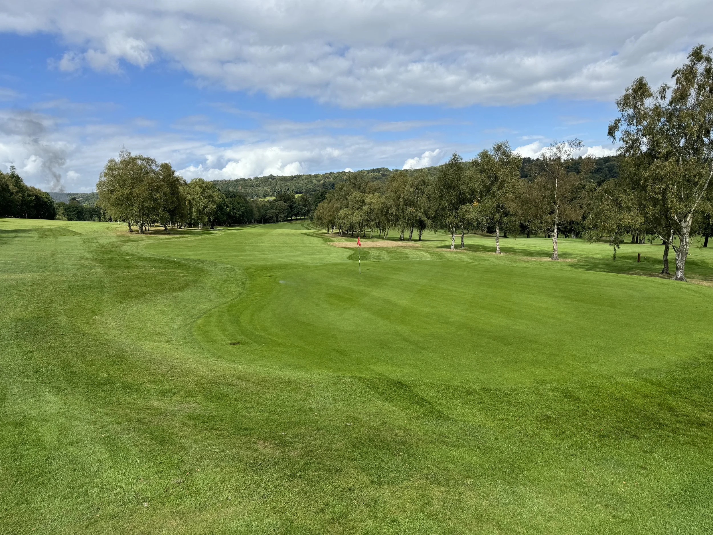 A golf course with green grass, sand traps, a red flag on the putting green, surrounded by trees, under a partly cloudy sky.