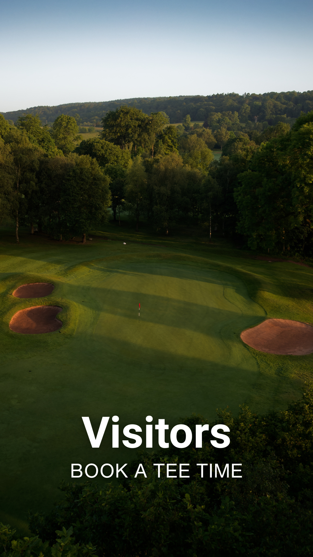 A scenic view of a golf course with green fairways, sand bunkers, and a flagstick, surrounded by trees and hills under a clear sky.