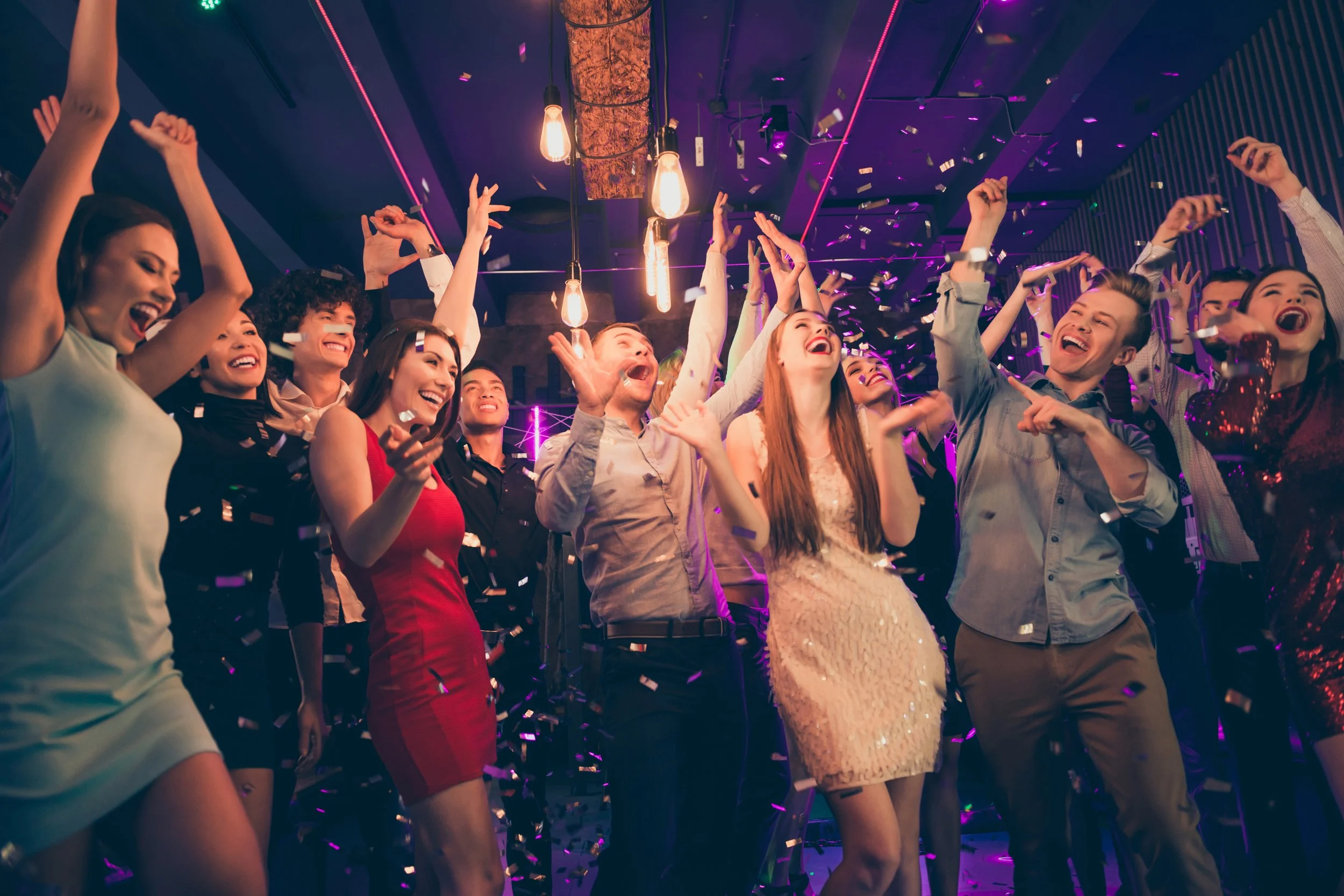 Group of people dancing and celebrating at a party with confetti and purple lights.