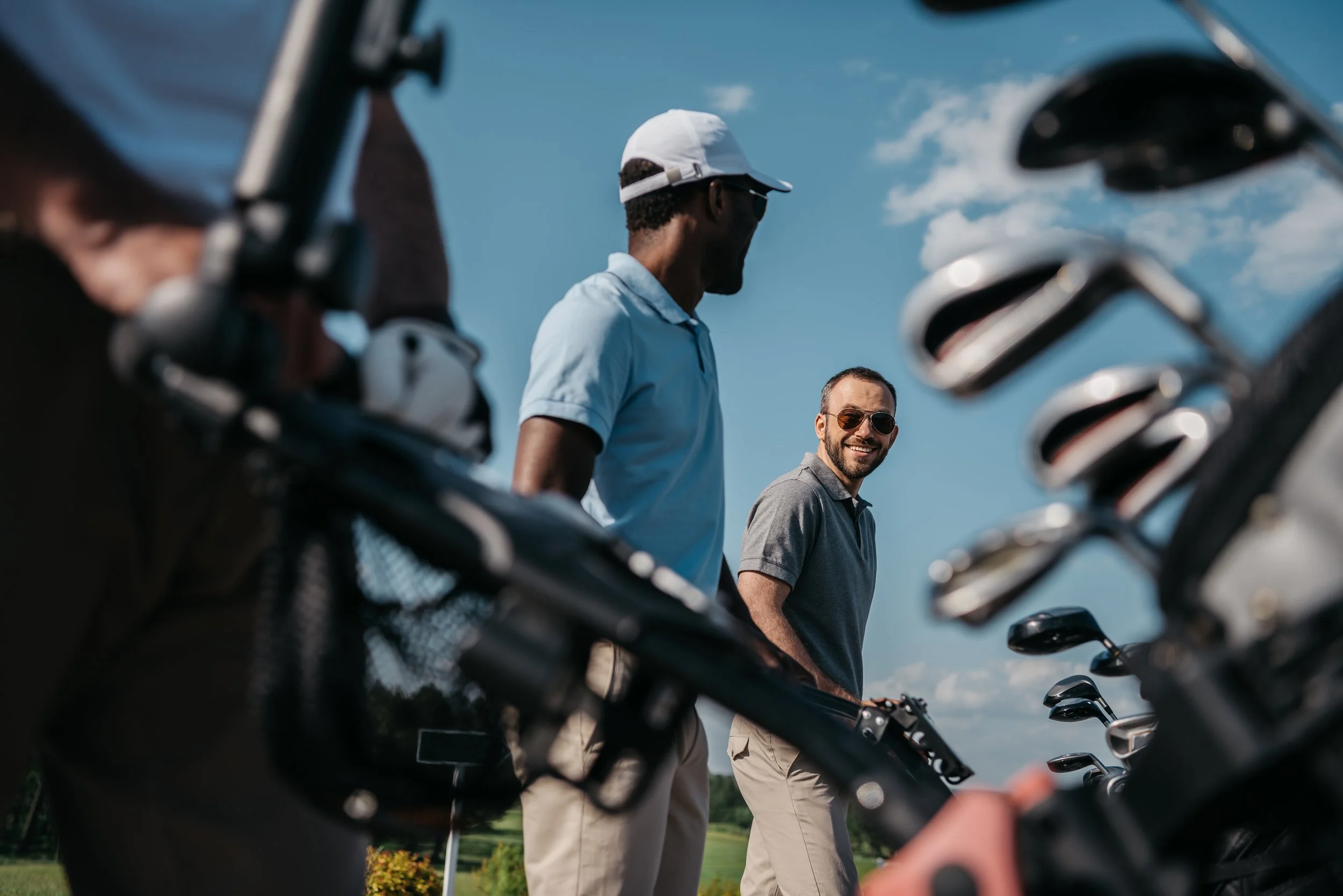 Two men standing outdoors near golf clubs on a golf course on a sunny day, with a blue sky and some clouds in the background.