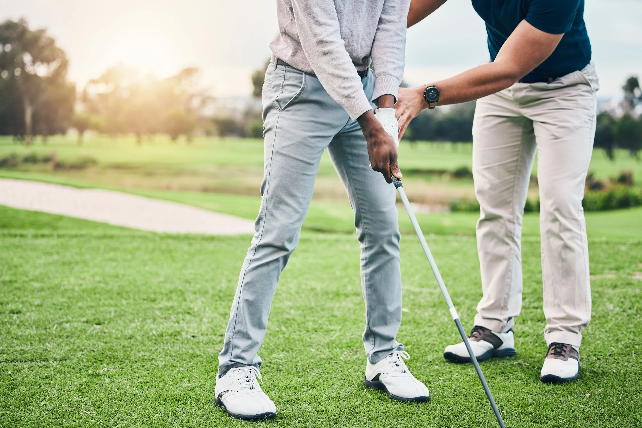 Two people on a golf course, one is helping the other with their golf swing. The person being helped is holding a golf club, and the other person is pointing or adjusting something on the club. The scene is outdoors with grass and trees in the background, under a bright, sunny sky.