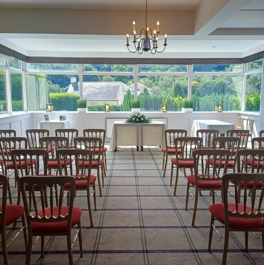 A beautifully decorated indoor event space with chairs arranged in rows facing a head table, which has a floral centerpiece. The room has large windows with a view of greenery and houses outside, letting in natural light. There is a chandelier hanging from the ceiling and small lanterns on the window ledge.