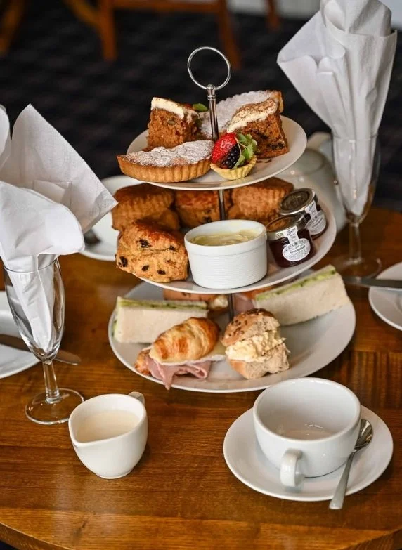 Afternoon tea set with assorted sandwiches, scones, pastries, jam, clotted cream, and a tiered stand of desserts, served on a wooden table with teacups, a teapot, and wine glasses with napkins.