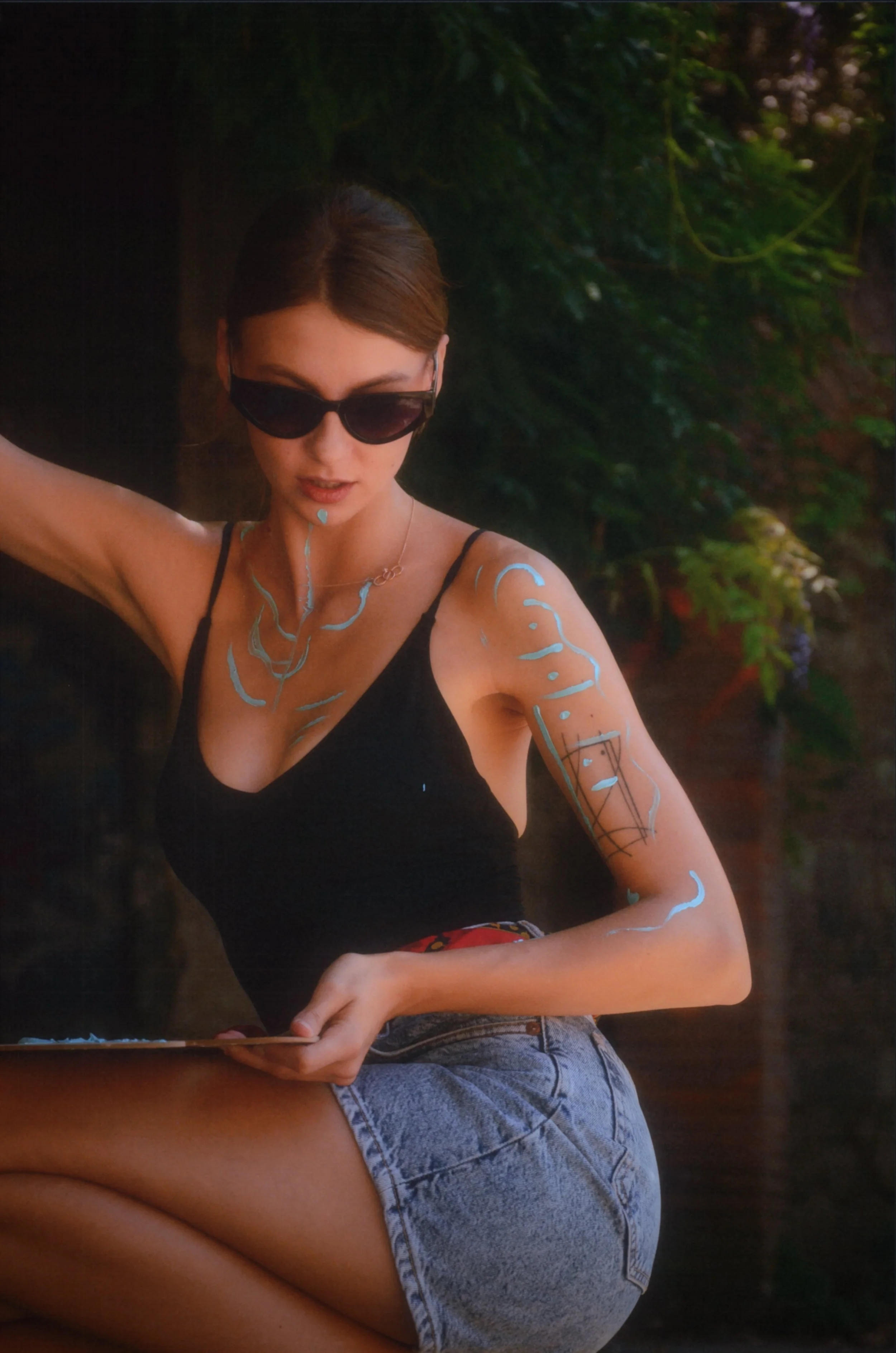 A woman with brown hair, wearing black sunglasses, a black tank top, and denim shorts, sitting outdoors near green foliage, with paint on her body.