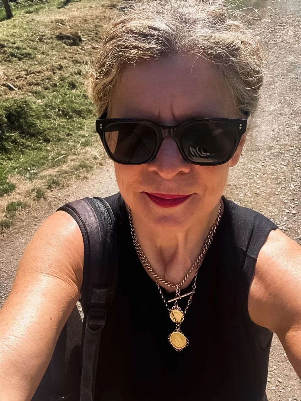 A person with gray hair wearing large black sunglasses, red lipstick, a black sleeveless top, a silver chain necklace with gold coins, and a backpack, standing on a dirt trail outdoors.