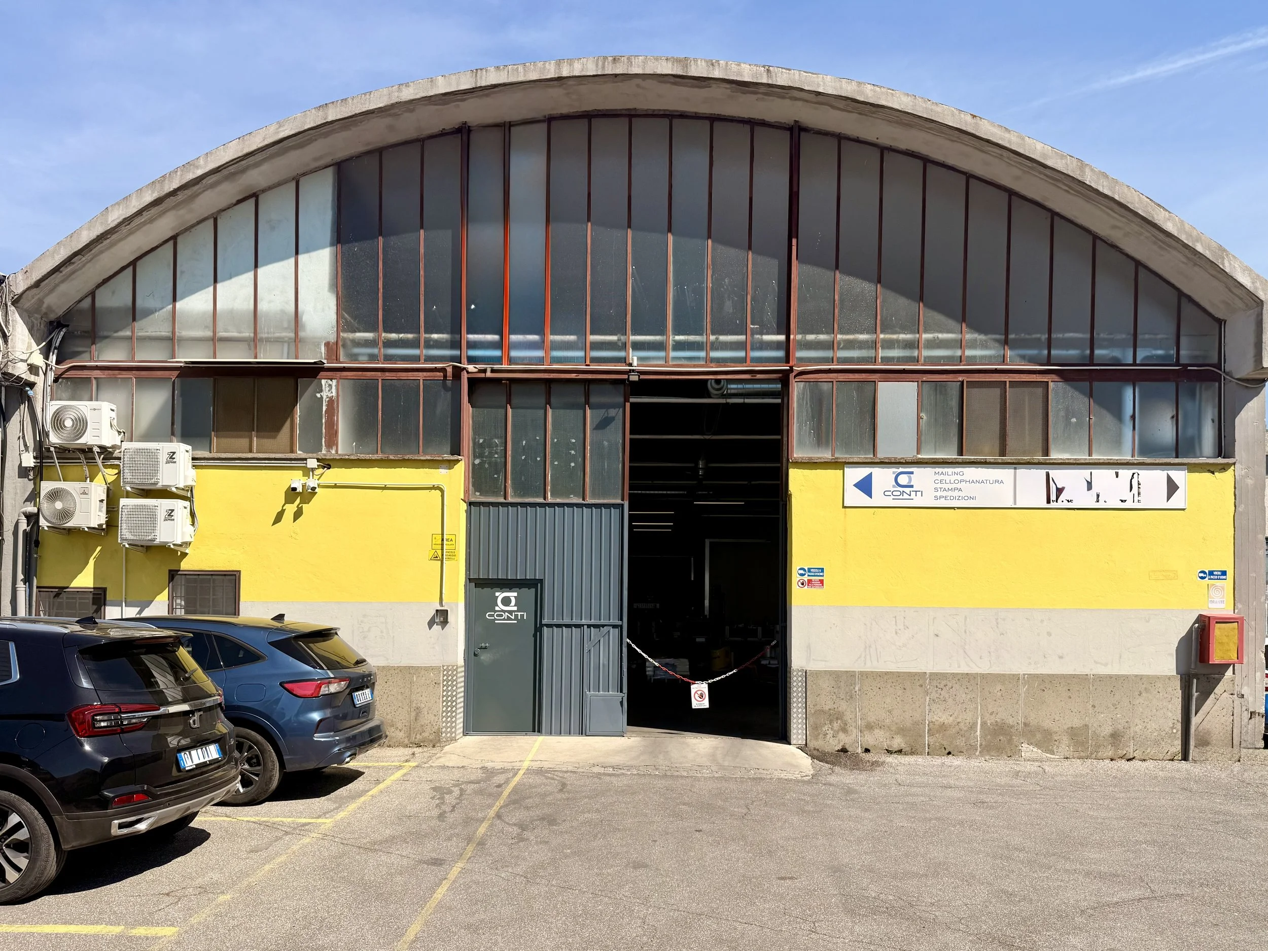 Facciata di un edificio commerciale con un grande finestrone curvo in vetro e una porta d'ingresso metallica. La parte inferiore è dipinta di giallo e presenta alcuni condizionatori d'aria installati sulla parete. In un cartello si vedono le parole 'CONT', e indicazioni per servizi come spedizioni e stampa.