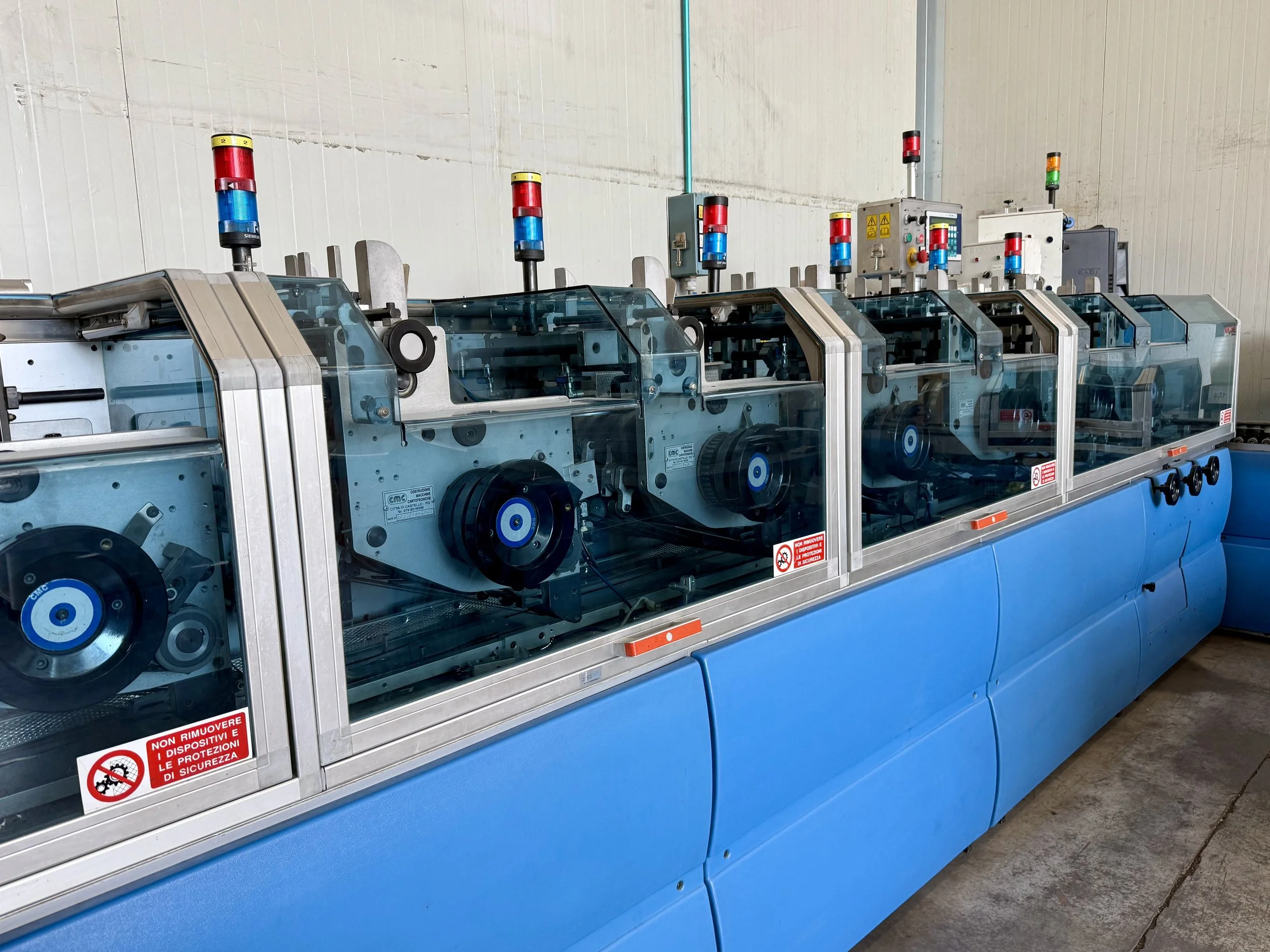 Macchine industriali con luci di segnalazione rosse e blu, protette da pannelli di vetro e situate in un ambiente industriale. Sono posizionate in fila su una superfice bluista, con un pannello di controllo e segnali di sicurezza visibili.