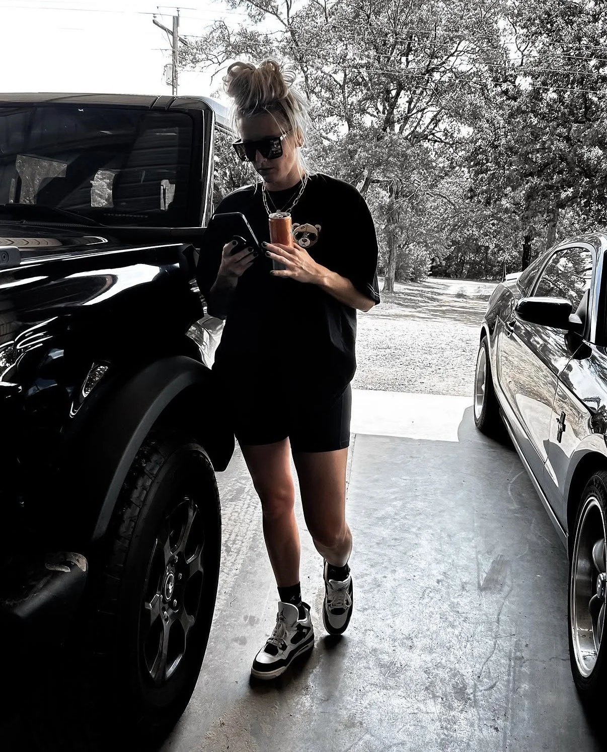 Woman wearing sunglasses, black t-shirt with bear graphic, shorts, and sneakers, standing next to black and silver cars, holding a smartphone and a drink, in a garage with trees and power lines in the background.