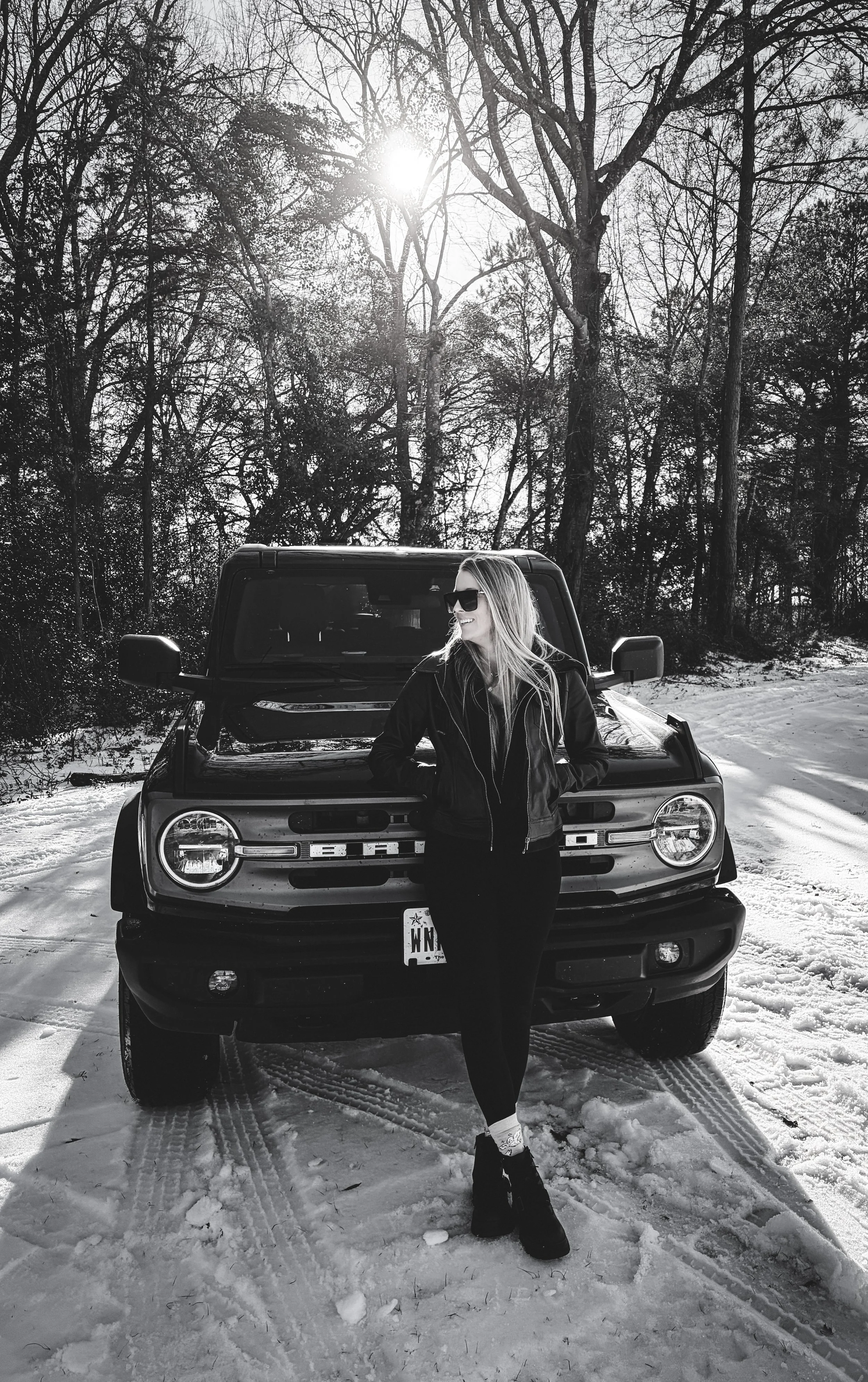 A woman with sunglasses and long hair smiling, standing in front of a black SUV on a snow-covered ground, surrounded by trees with no leaves and a bright sun in the sky.