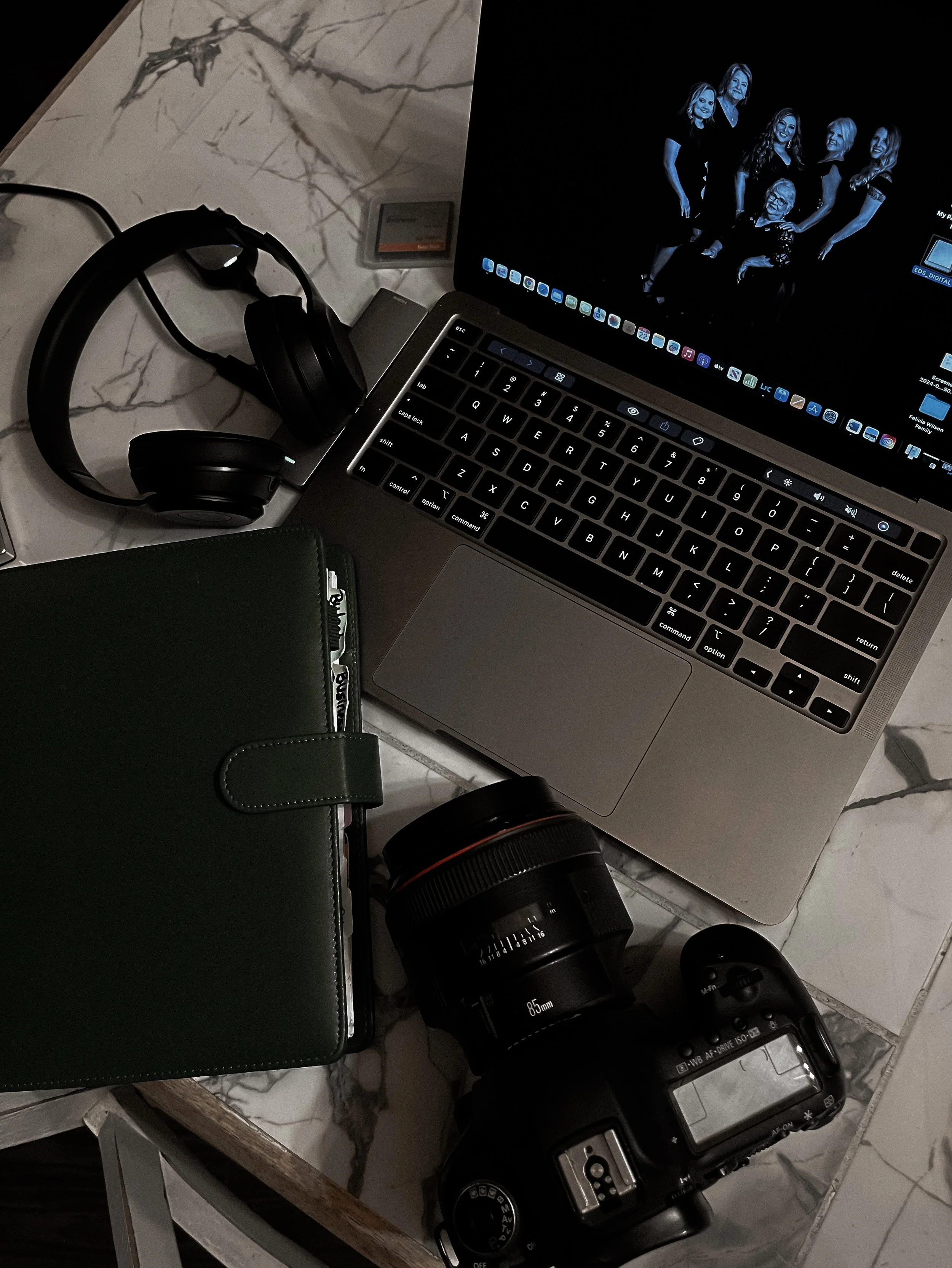 A laptop, a pair of black headphones, a camera lens, a camera body, a closed green notebook, and a piece of paper on a marble table.