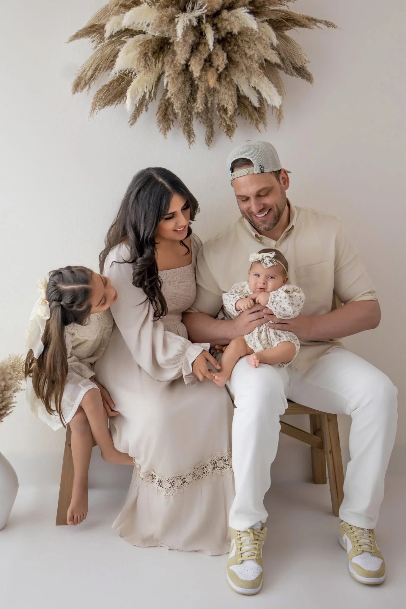 A family of four, including two young girls and their parents, sitting on a white bench against a white wall decorated with a large dried pampas grass arrangement. The mother and one of the girls are dressed in light-colored clothing, and the father is wearing a beige shirt, a backward gray cap, and white pants. The baby girl is dressed in a patterned outfit with a bow headband, and the family appears to be smiling and happy.