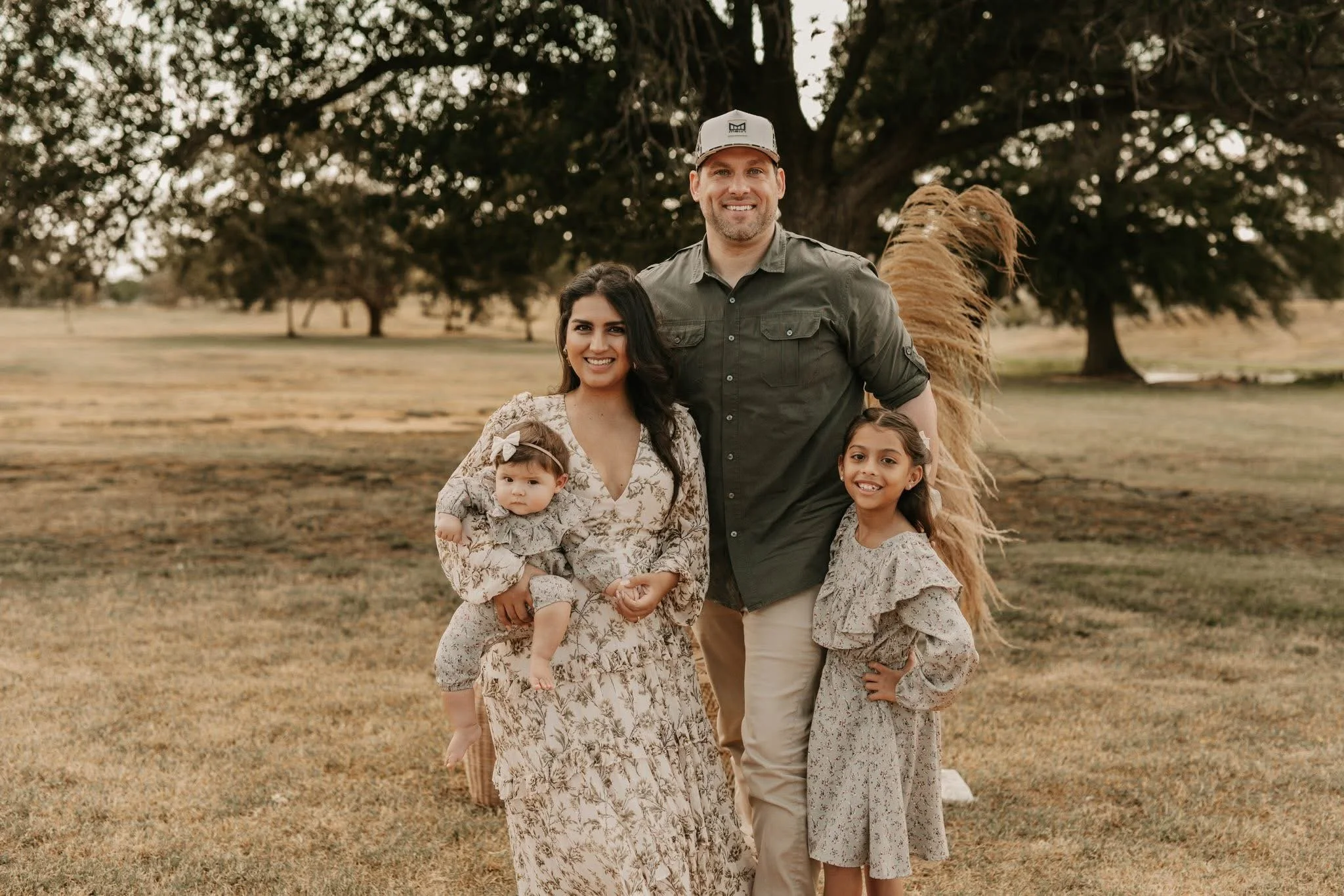 A smiling family of four, including a mother, father, and two young daughters, standing outside in a park with large trees and dry grass, during sunset.