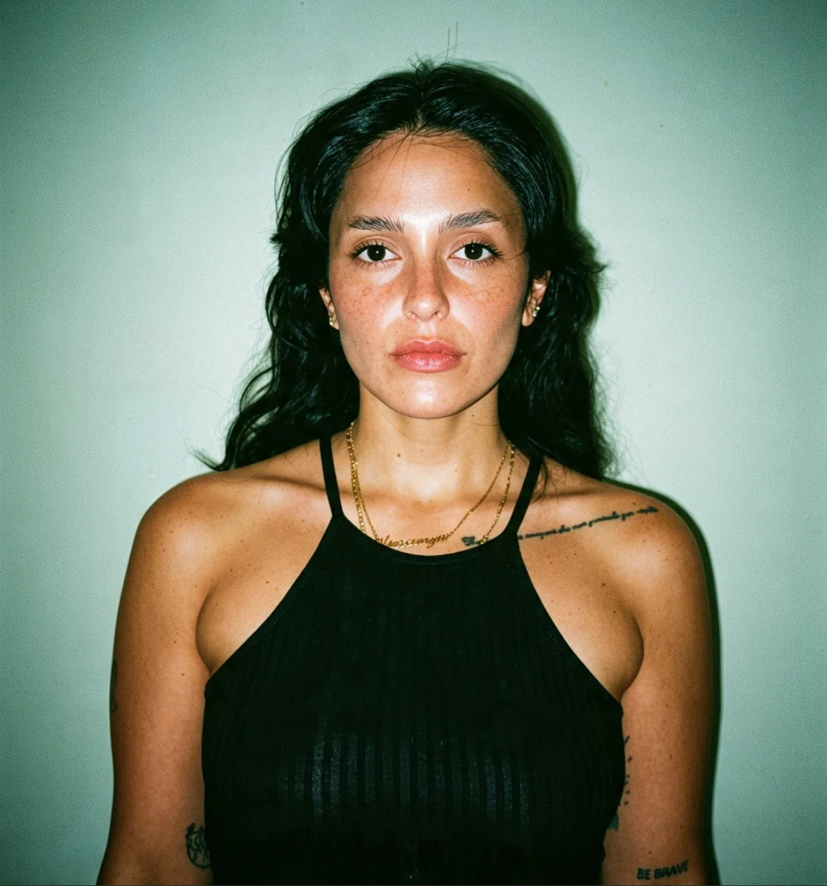A woman with long dark hair and freckles, wearing a black tank top, gold necklaces, and earrings, standing against a plain light-colored background.