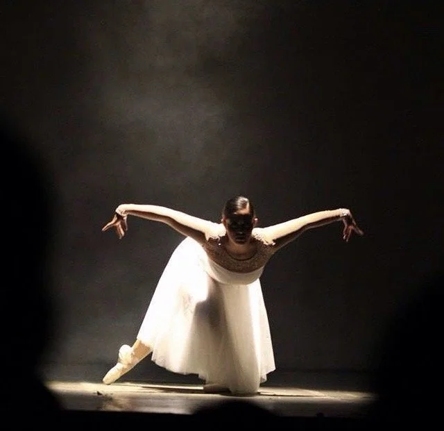 Ballet dancer in a white tutu bowing on stage with her arms extended, in a dimly lit setting.