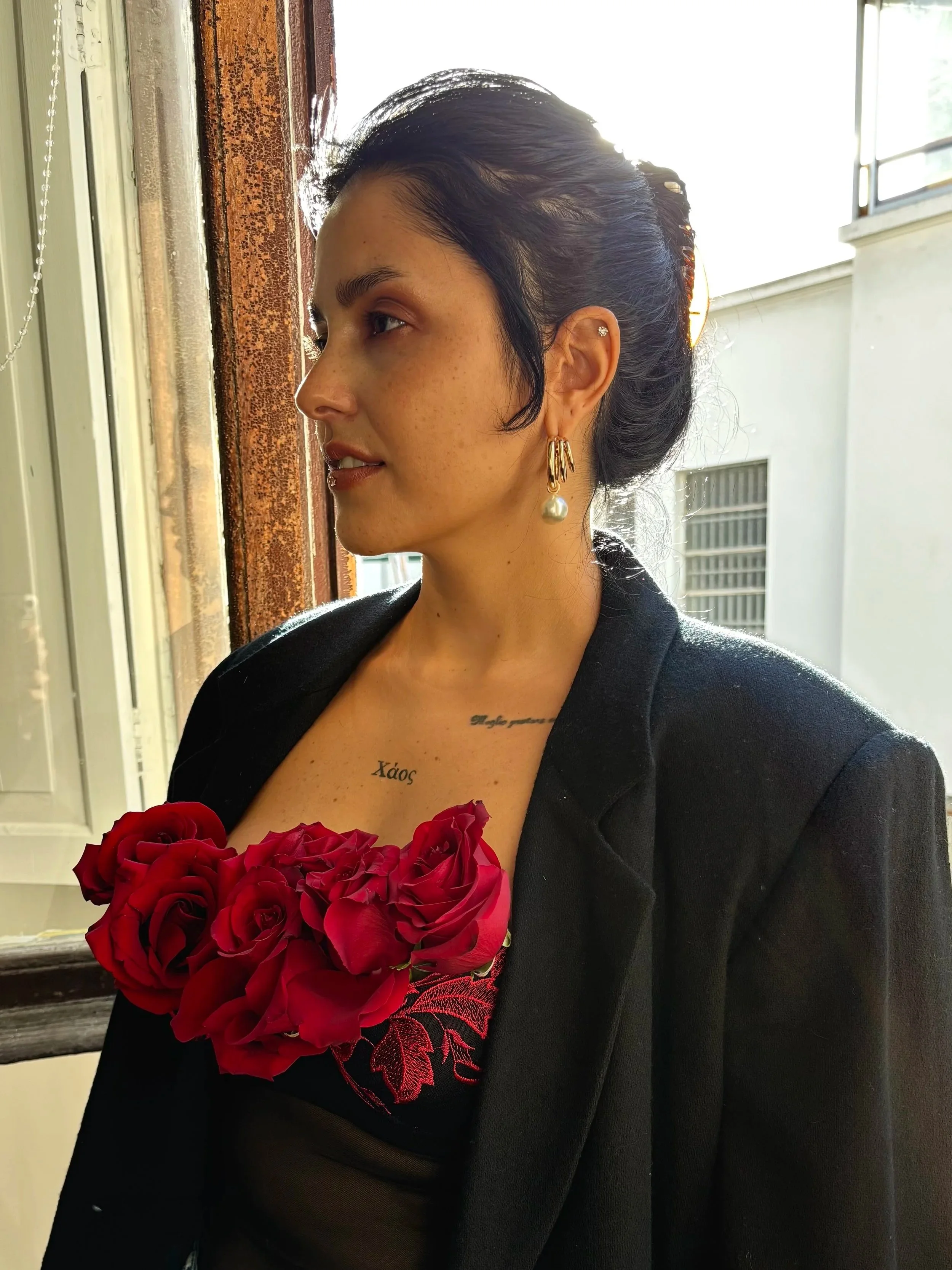 A woman with black hair styled in an updo, wearing large gold hoop earrings with pearls, holding a bouquet of red roses, and wearing a black blazer with a visible tattoo on her chest.