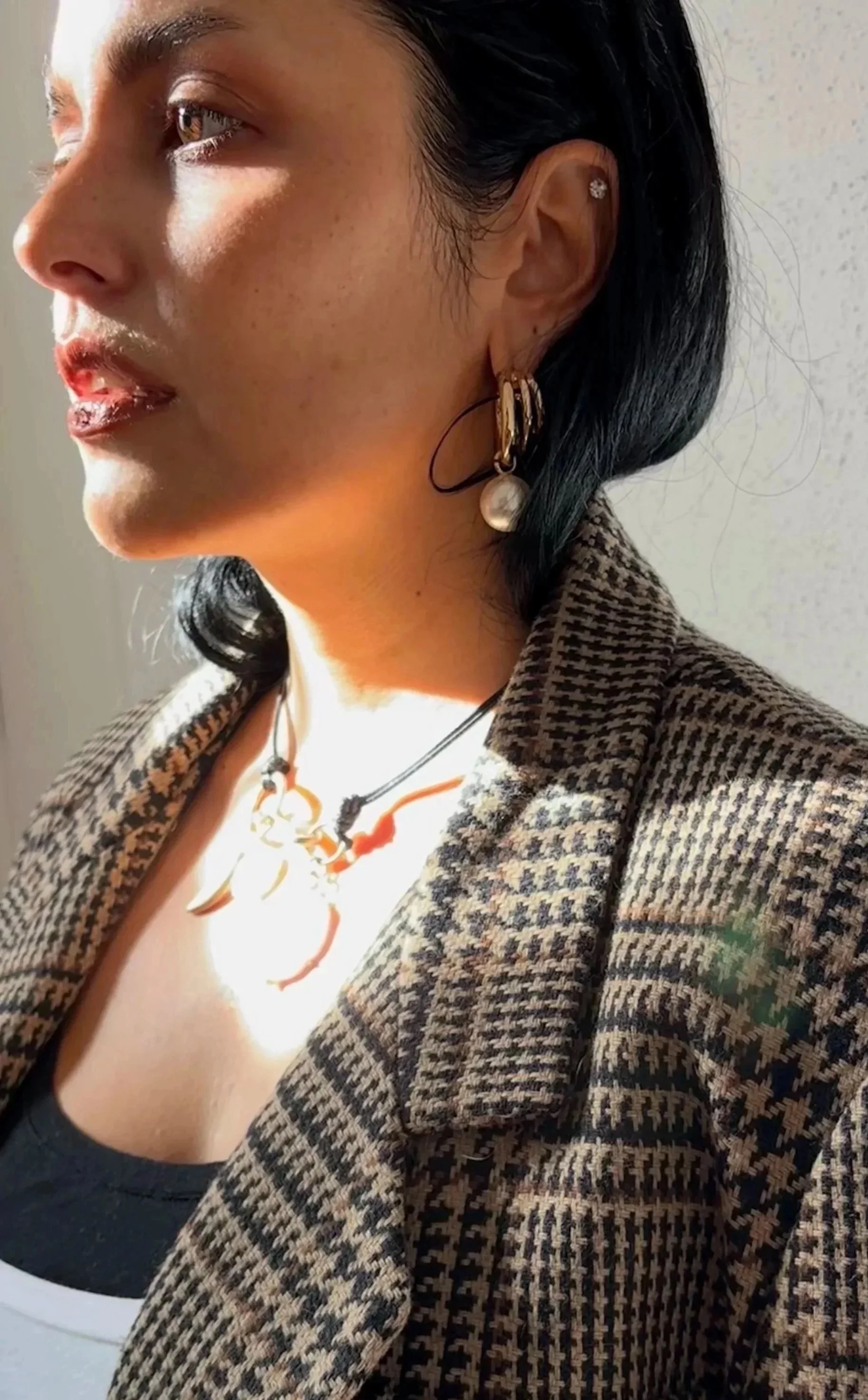 Close-up of a woman with dark hair wearing gold hoop earrings, a pearl earring, layered necklaces, a checkered blazer, and a black top, with sunlight illuminating her face.