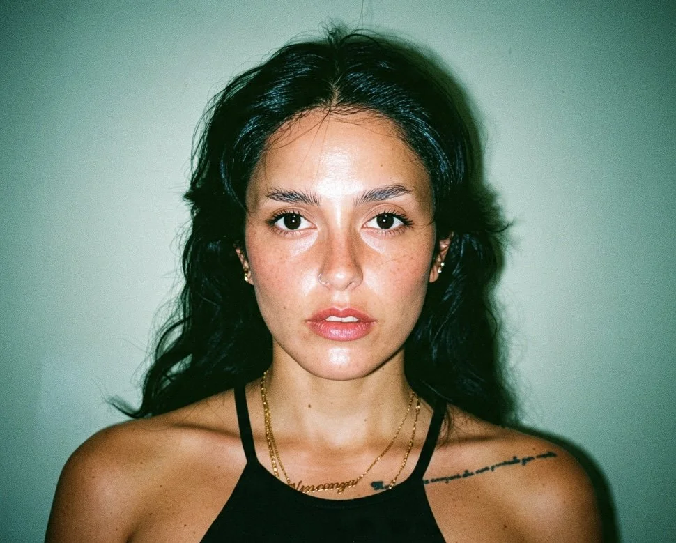 A young woman with long dark hair, wearing a black tank top, gold necklaces, and jewelry, looking directly at the camera with a neutral expression, against a plain light background.