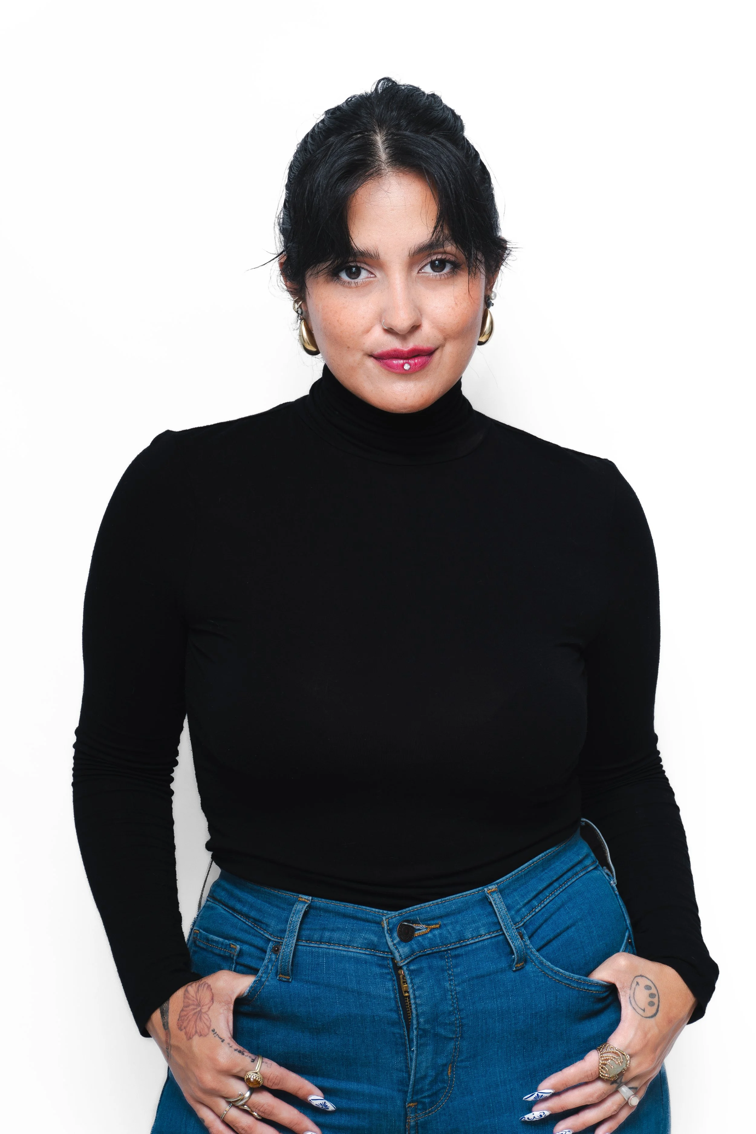 A woman with black hair, wearing a black turtleneck shirt and blue jeans, is standing against a white background. She has tattoos on her hands and fingers, gold earrings, and a facial piercing. She is looking directly at the camera with a slight smil