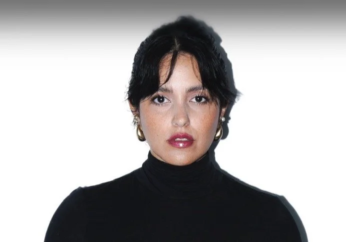 A woman with short black hair wearing a black turtleneck and gold earrings, looking directly at the camera with a neutral expression.