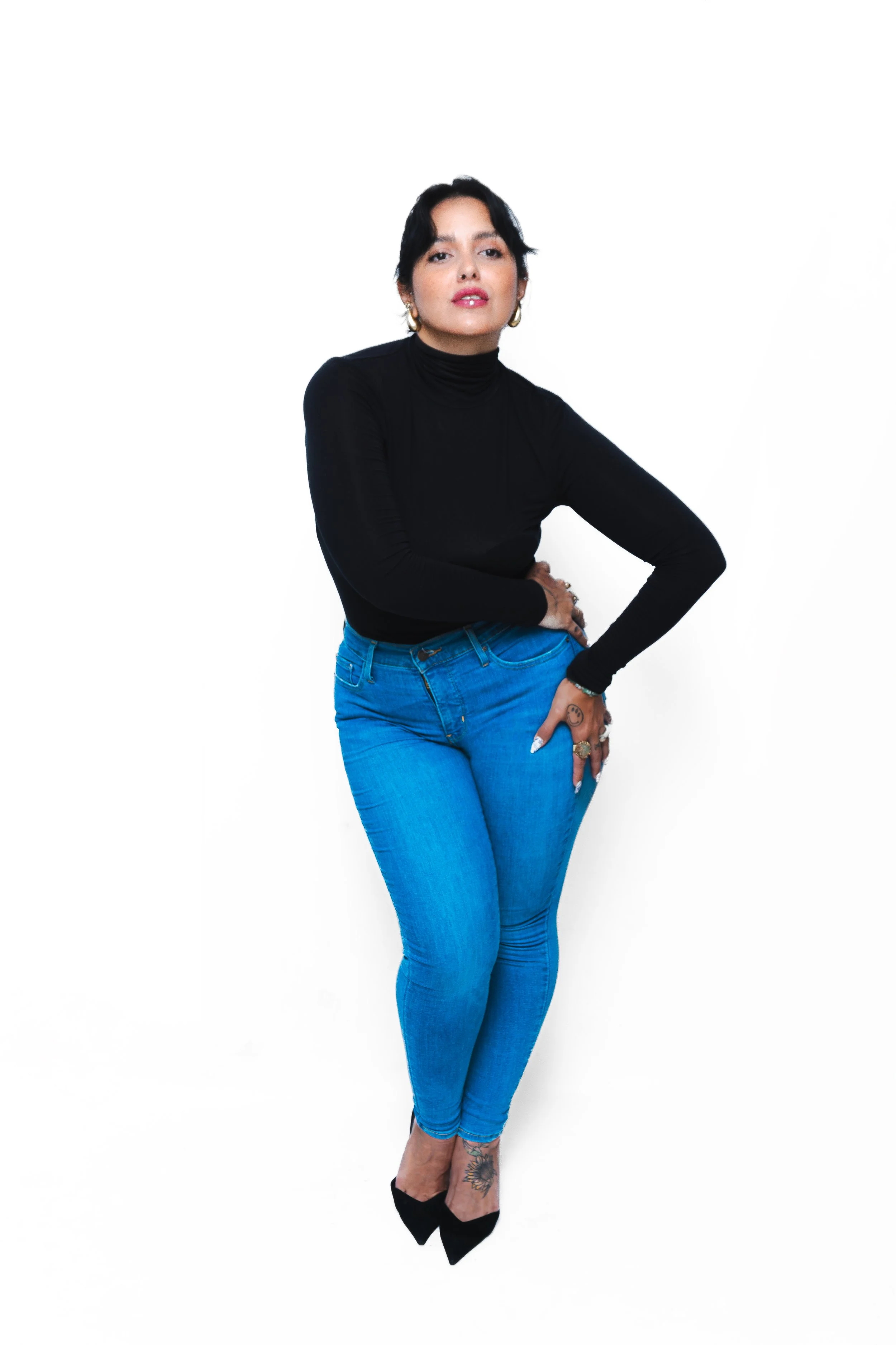 A woman with short dark hair and hoop earrings wears a black turtleneck and blue jeans, standing with one hand on her hip and the other hanging down, against a white background.