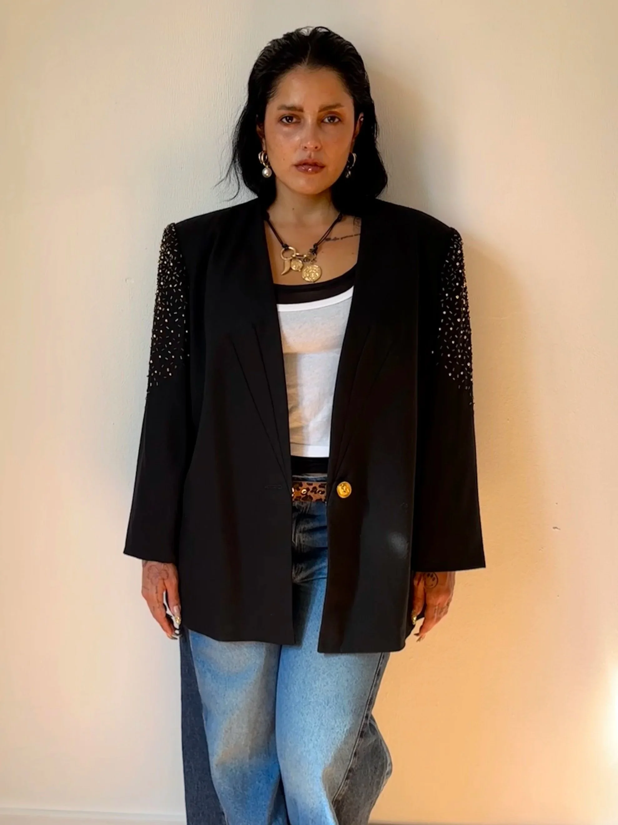 A woman with dark hair, wearing a black blazer with sparkly sleeves, turquoise jeans, and layered necklaces, standing against a plain wall.