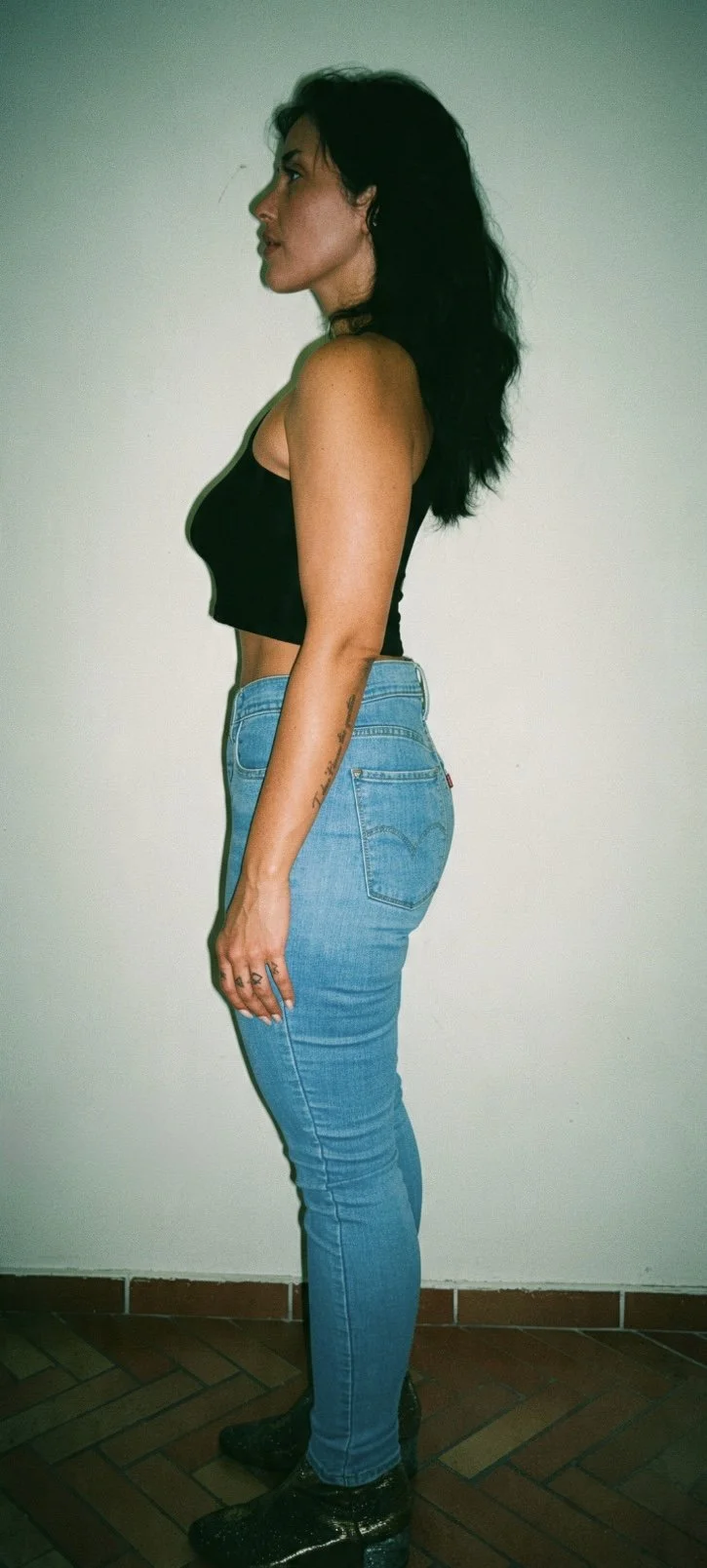 Side profile of a woman with dark hair, black cropped top, blue jeans, and black boots standing against a plain wall.