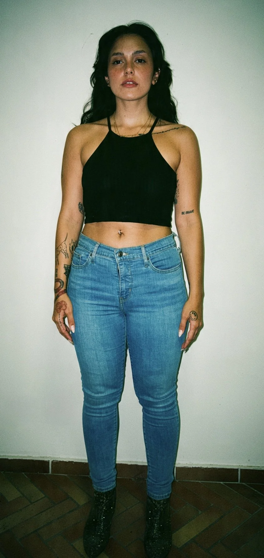 A woman standing against a plain white wall, wearing a black crop top, blue jeans, and black boots with glitter. She has long dark hair, multiple tattoos on her arms and torso, and is looking directly at the camera.