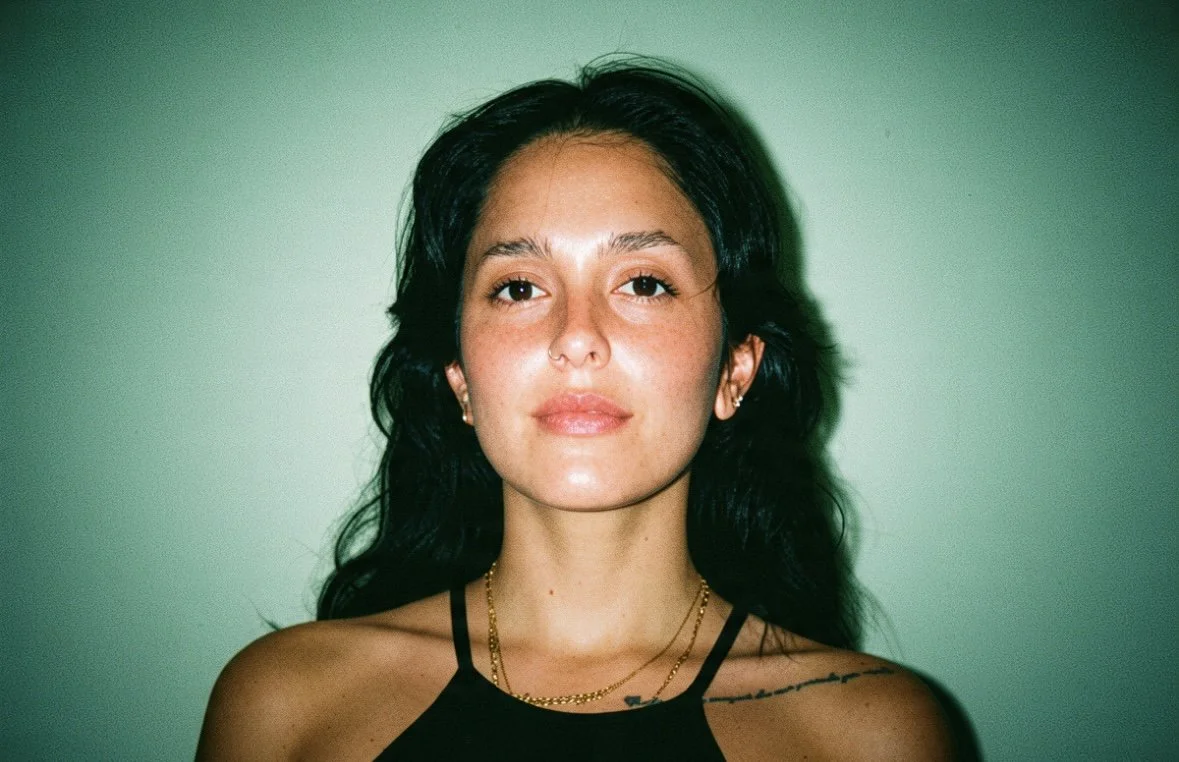 A woman with long dark hair, wearing a black tank top, gold necklaces, and earrings, standing against a green wall.