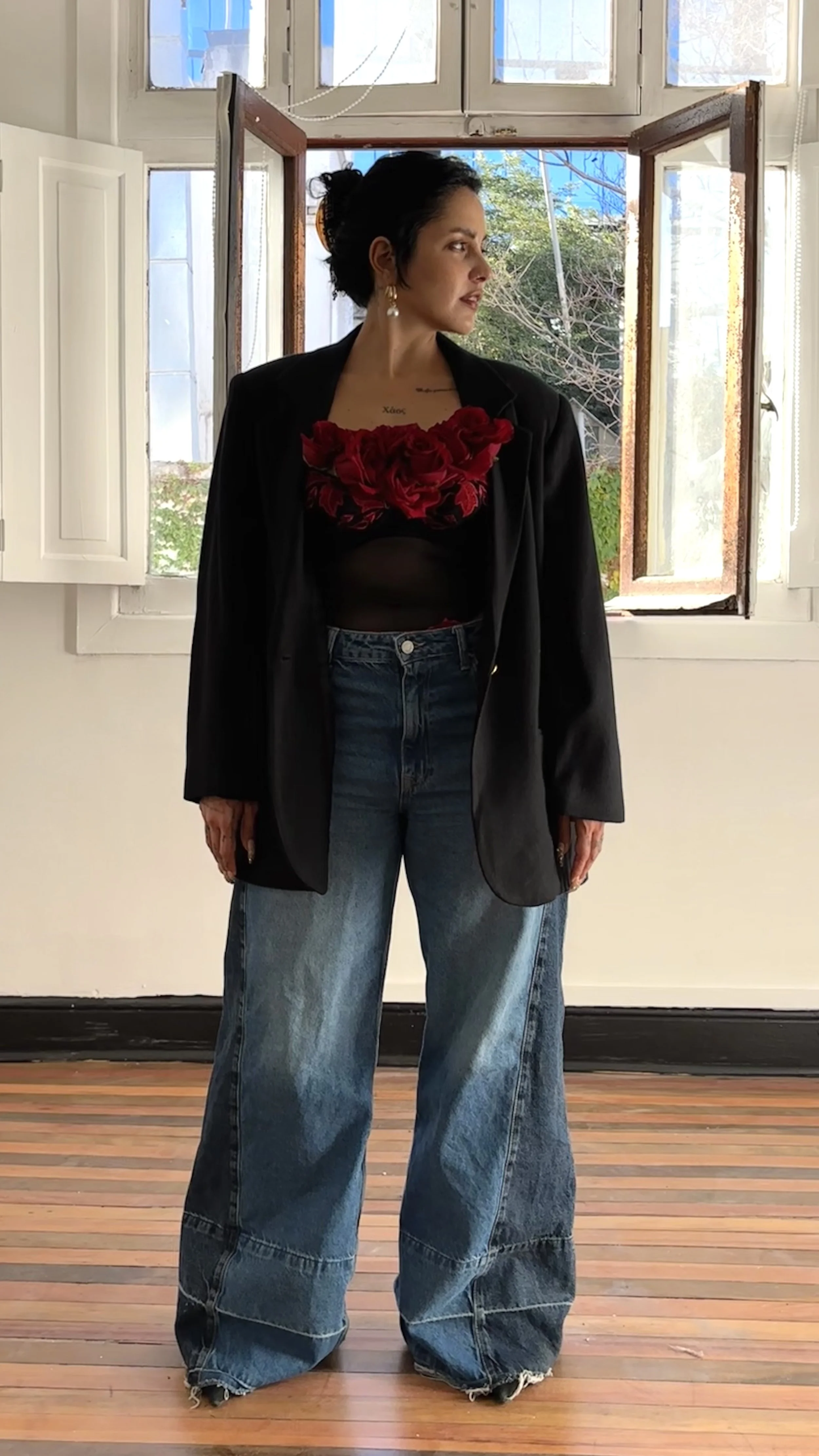 A woman standing indoors in front of an open window, wearing a black blazer, a sheer black top with red floral accents at the chest, and wide-leg blue jeans. She has dark hair styled in an updo, earrings, and a tattoo on her chest.