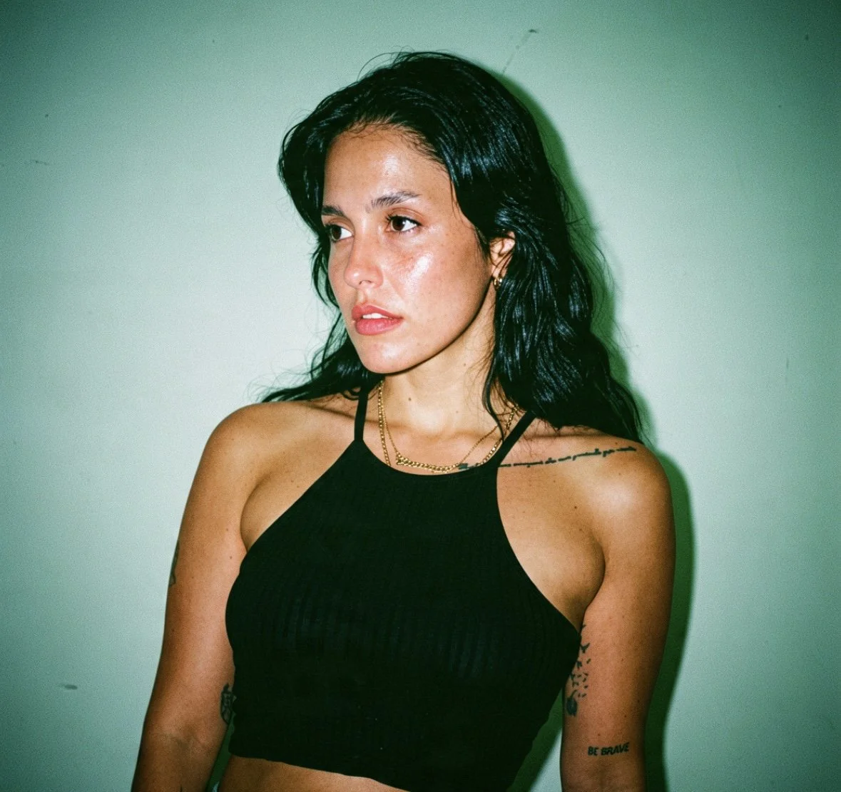 A woman with long black hair wearing a black tank top, gold jewelry, and tattoos on her arm and shoulder, standing against a plain wall.