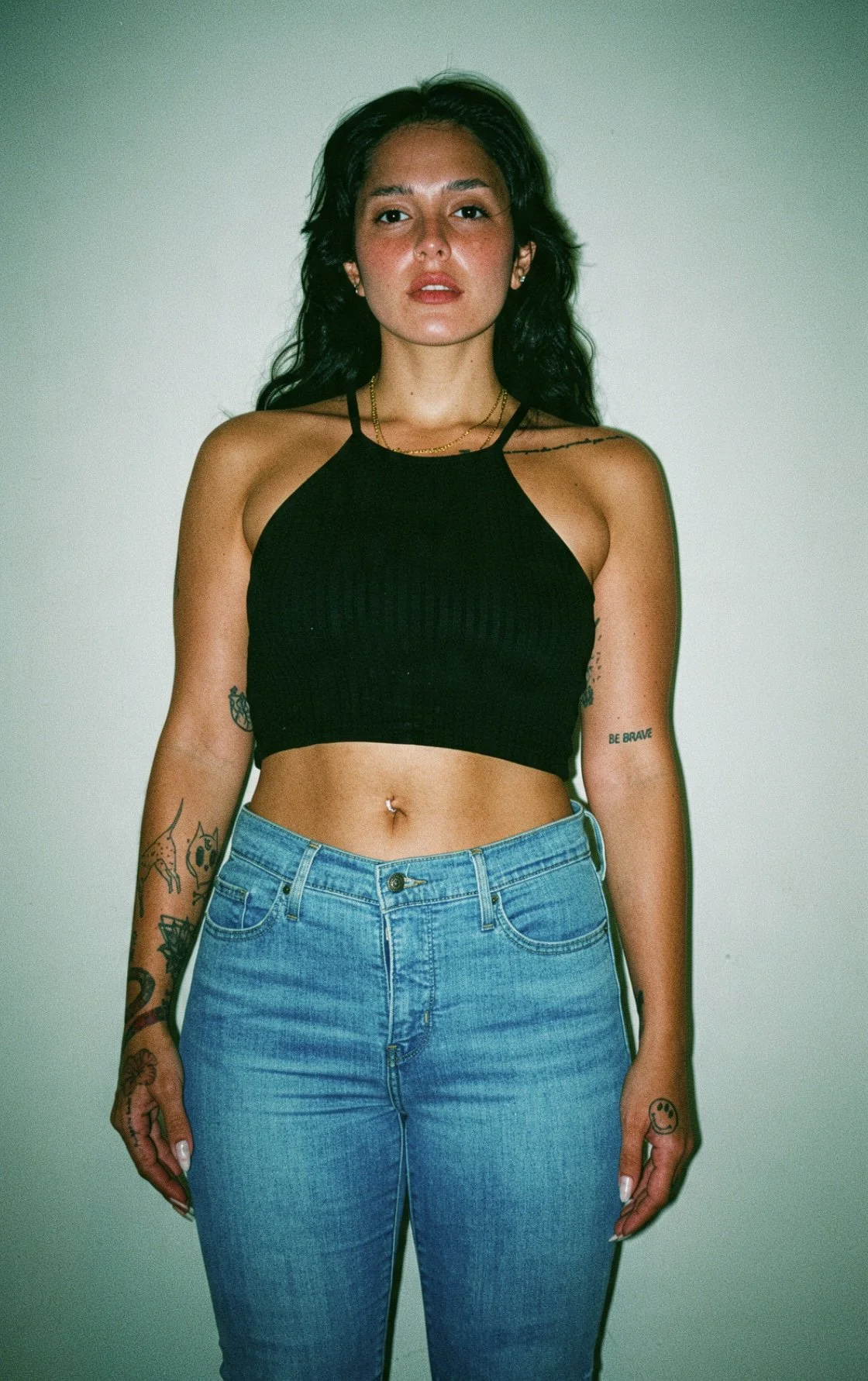 A woman with black hair and tattoos on her arms is wearing a black crop top and blue jeans, standing against a plain background.