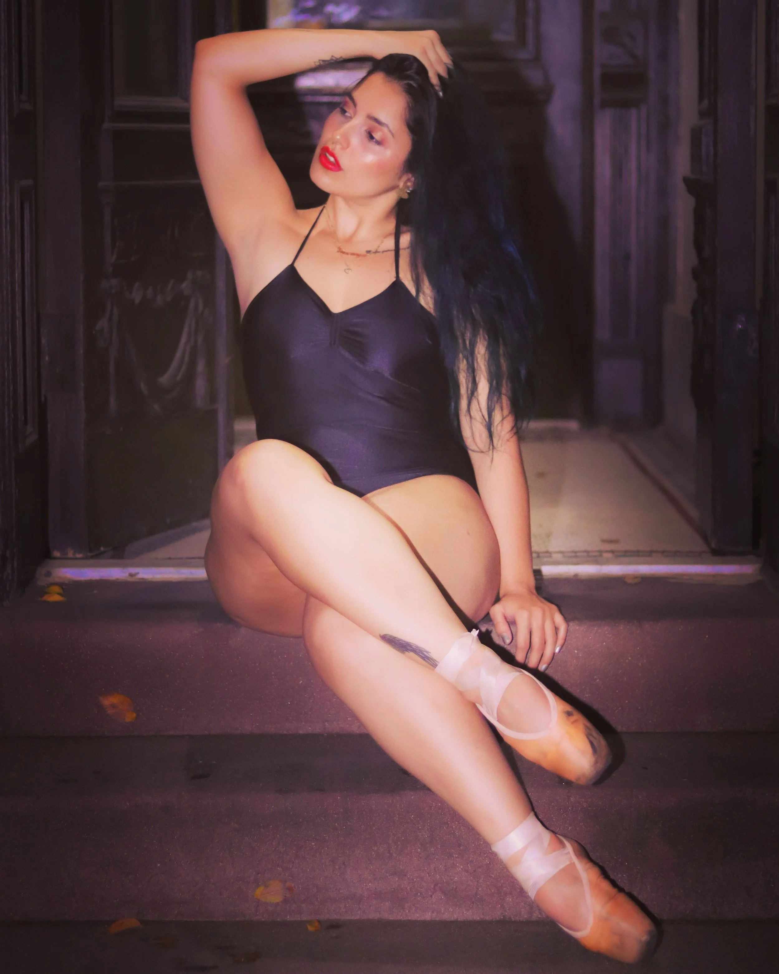 A woman in a ballet pose, wearing a black dress and ballet shoes, with one leg raised and wrapped in ballet tape, standing outdoors at night.