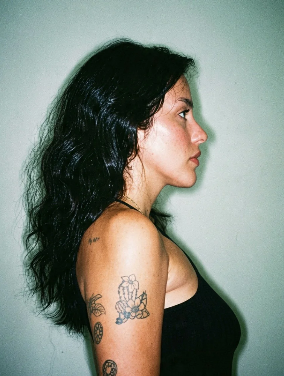 Profile of a young woman with dark, wavy hair and tattoos on her arm, wearing a black sleeveless top, standing against a plain light green wall.