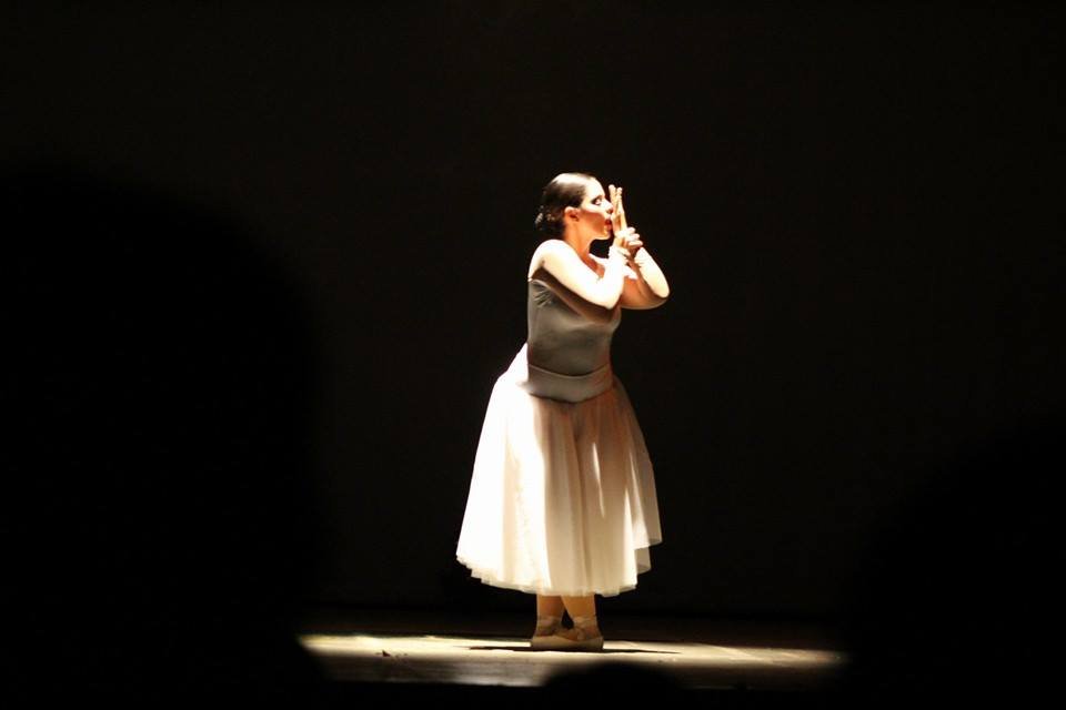 A woman walking on stage illuminated by a spotlight, wearing a black top and a beige tulle skirt.