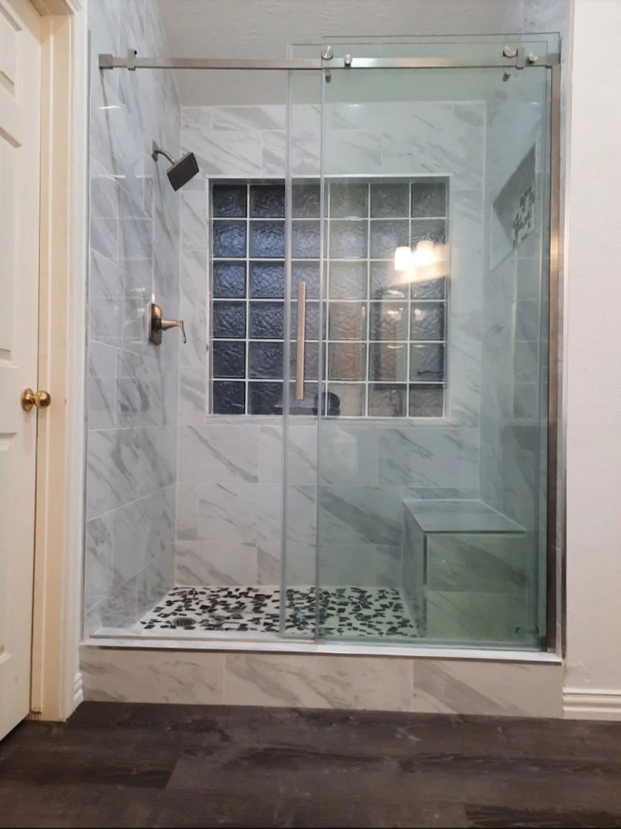 A modern glass-enclosed shower with a metal sliding door, marble walls, a small window made of textured glass blocks, a built-in bench, and a black and white pebble tile floor inside the shower. There is a wall-mounted showerhead and a separate contr
