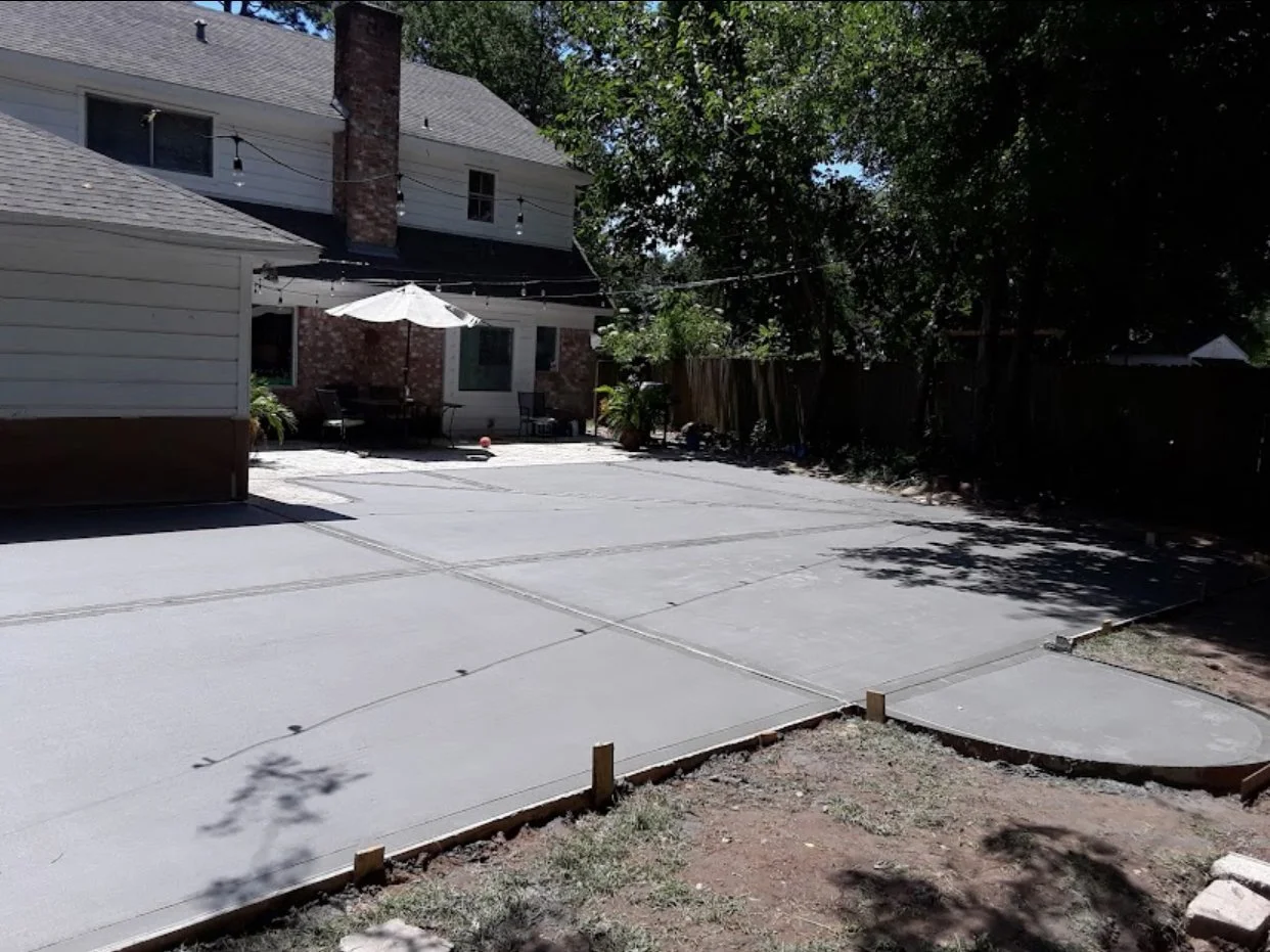 Backyard with newly poured concrete patio, outdoor furniture, and string lights, surrounded by trees and a house with a brick chimney.