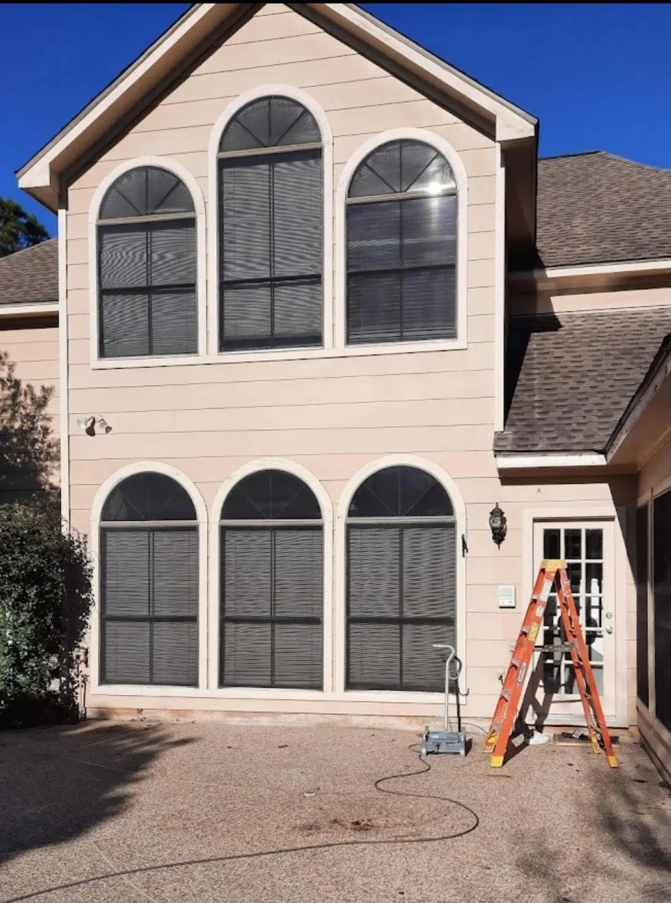 The exterior of a two-story house with peach siding and large arched windows, a ladder outside the door, and patio equipment on the ground under a clear blue sky.