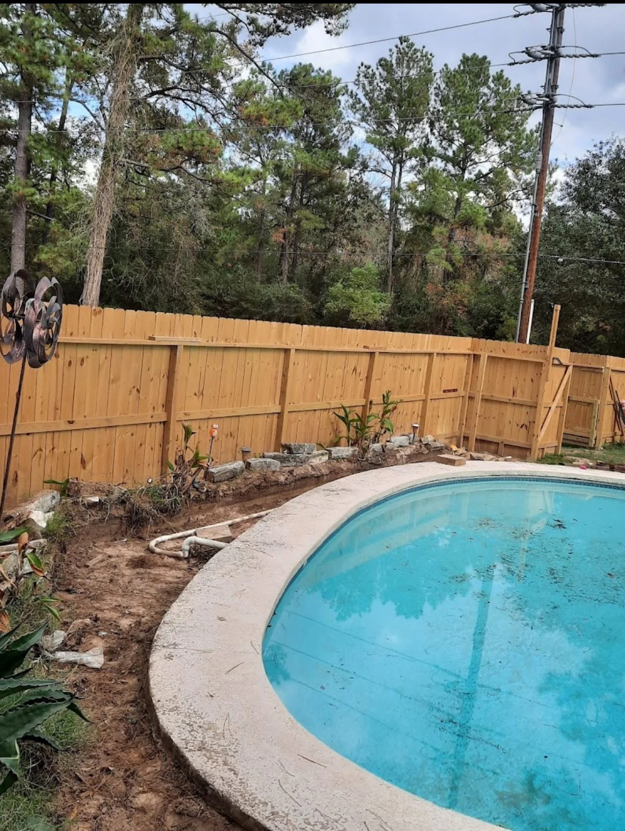 Backyard with a new wooden privacy fence and a swimming pool. The pool is partially filled with blue water, and there is construction work around the pool with soil, rocks, and a hose visible.
