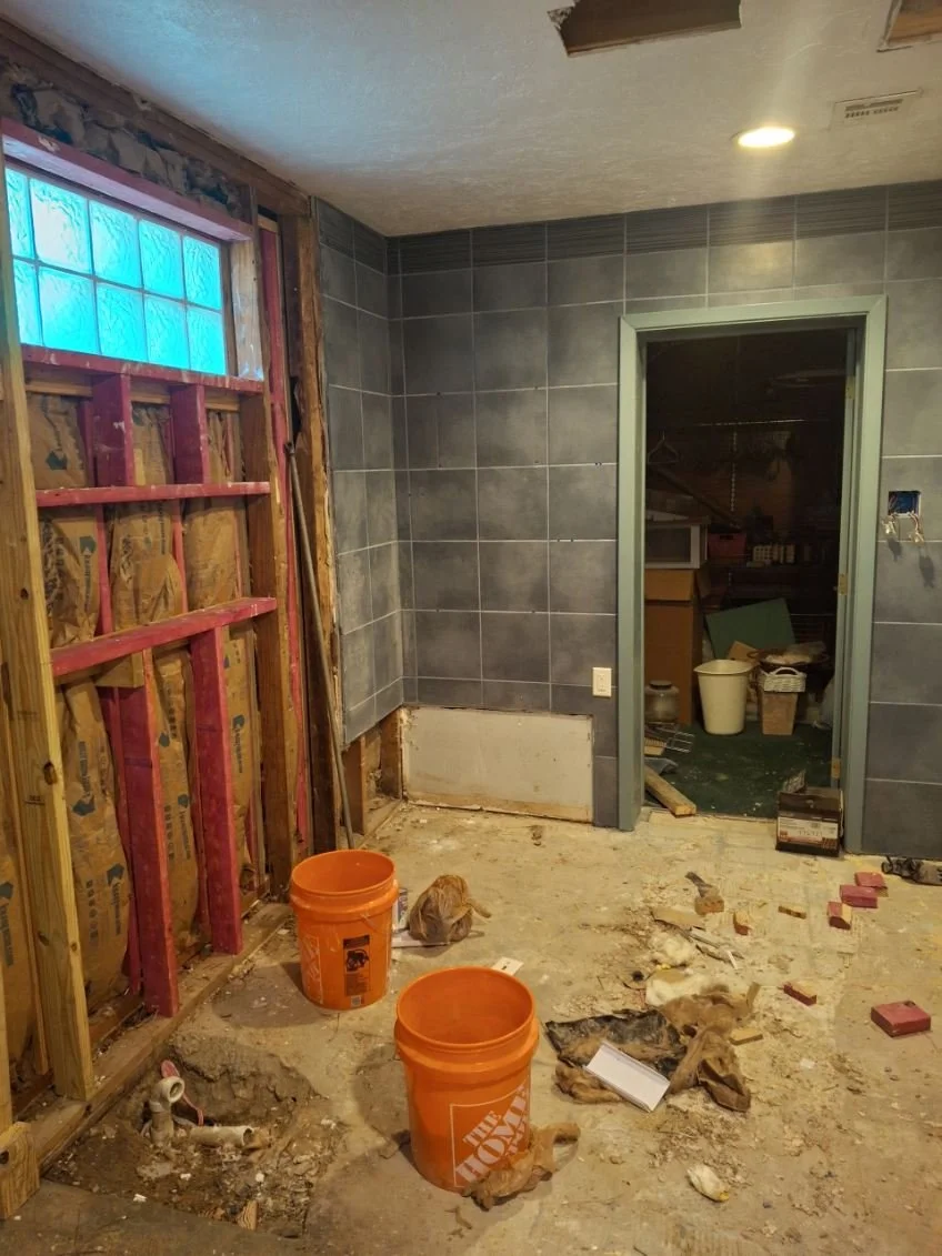 Bathroom under renovation with exposed wall studs, construction debris, two orange buckets, and a partially tiled wall.