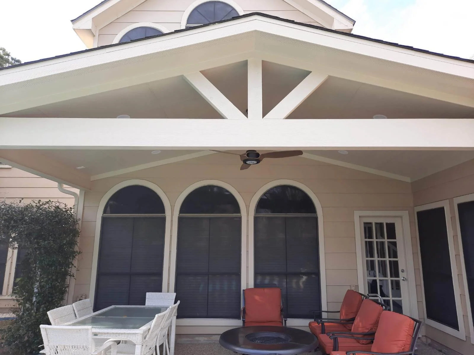 Back porch of a house with outdoor furniture including white chairs and orange cushioned chairs, a round fire pit, and large arched windows, with a beige exterior and a ceiling fan overhead.