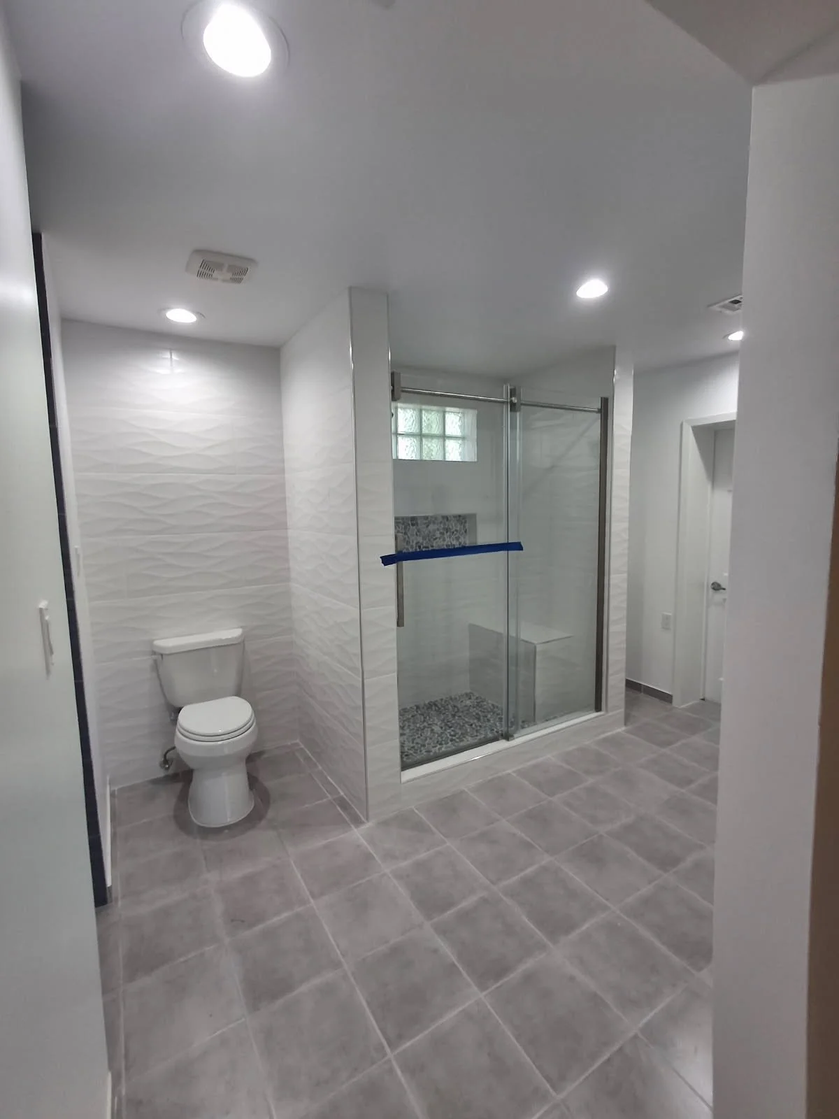 Modern bathroom with walk-in shower and toilet, white textured wall tiles, gray floor tiles, and recessed ceiling lights.