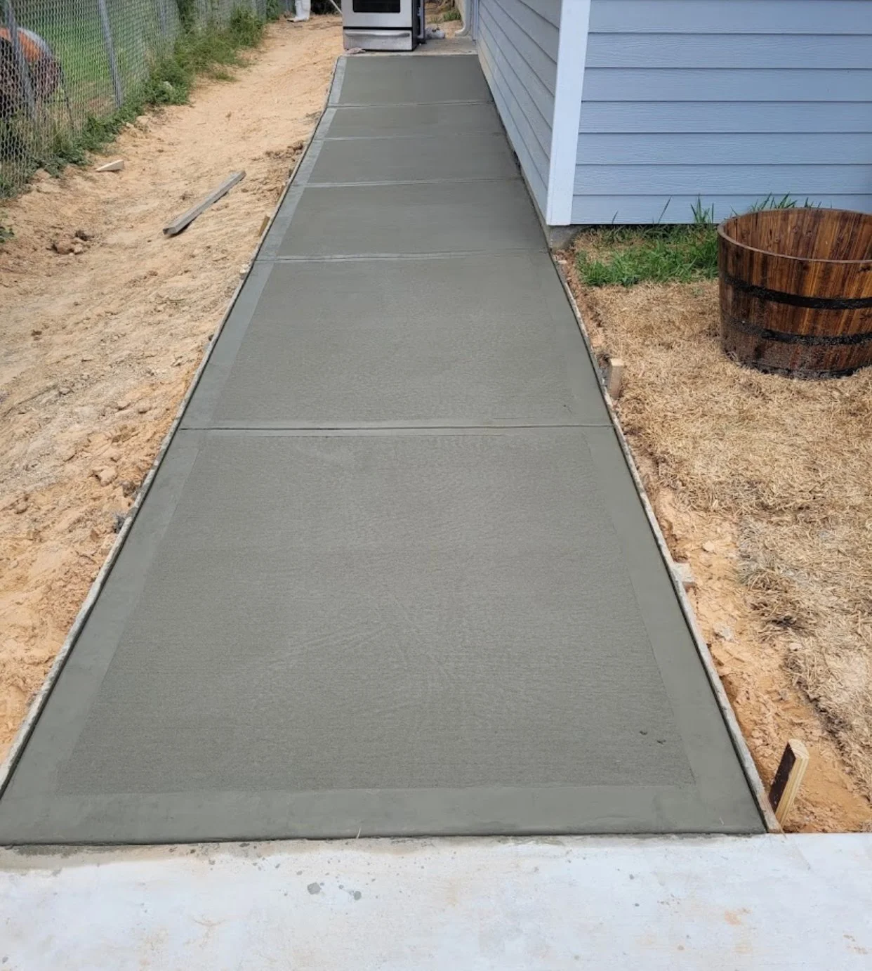 Freshly poured concrete sidewalk outside a house, bordered by a metal frame, with a gray exterior wall nearby and a wooden planter to the right.