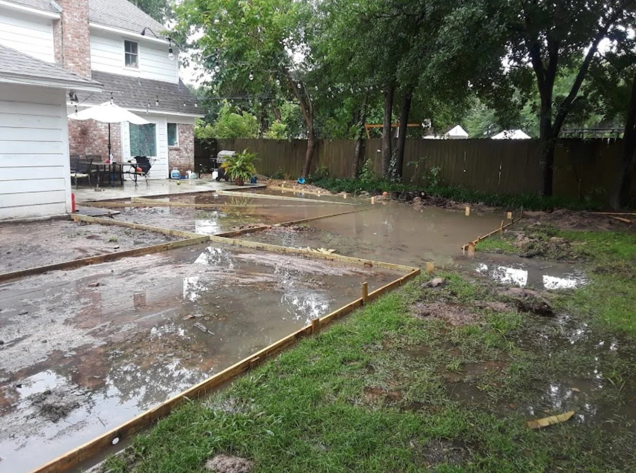 A backyard with a wet concrete slab, which is being prepared for a patio or new surface, with some parts flooded with rainwater, surrounded by a grassy area, trees, and a wooden fence, and a house with outdoor furniture and string lights in the backg