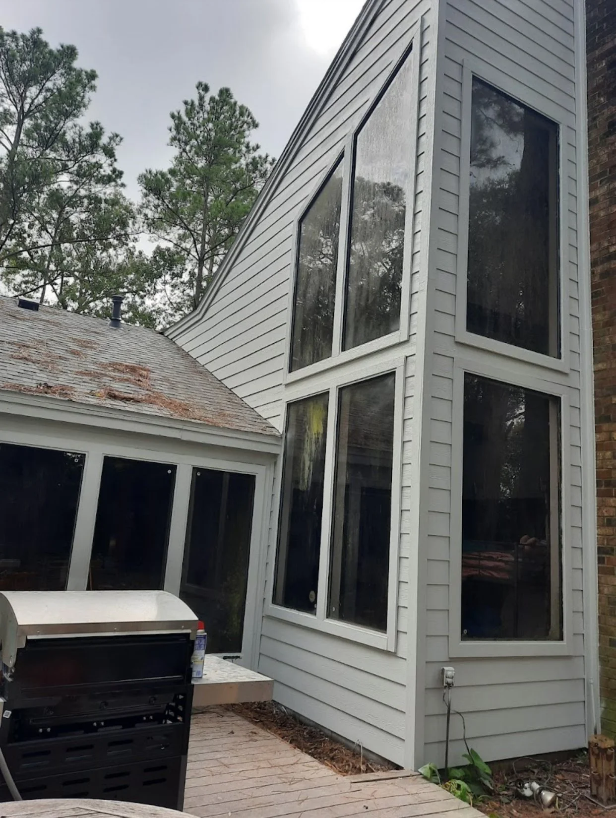 A two-story house with multiple large windows in white window frames and siding. The roof appears aged with some rust stains. There's a small patio with a grill and some patio furniture visible in the foreground. Tall trees are in the background.