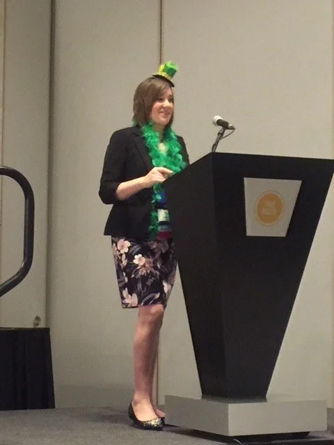 A woman standing at a podium, wearing a black blazer, floral skirt, and a small green hat, with a green feather boa around her neck, speaking or presenting at an event.