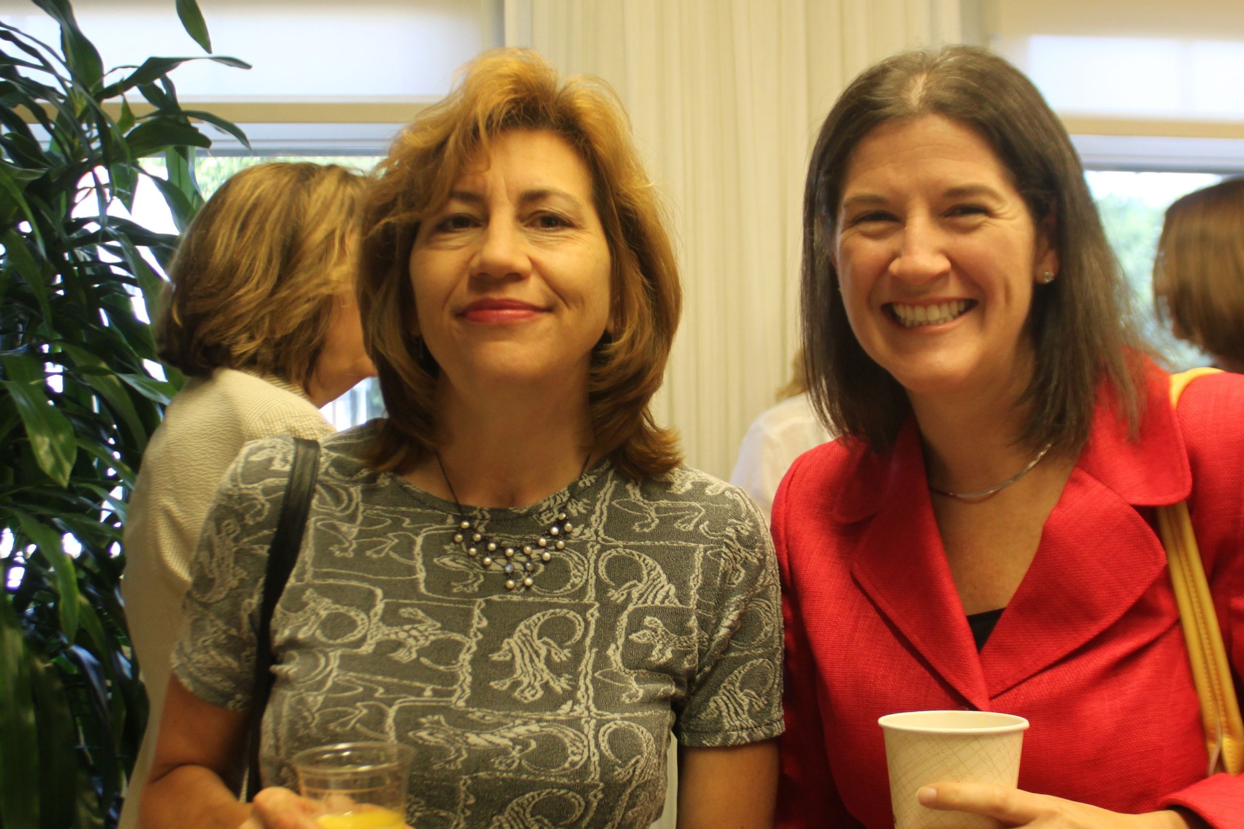 Two women smiling at an indoor gathering, holding drinks in paper cups. One woman has shoulder-length brown hair and is wearing a patterned top, the other has dark brown hair and is wearing a red blazer.