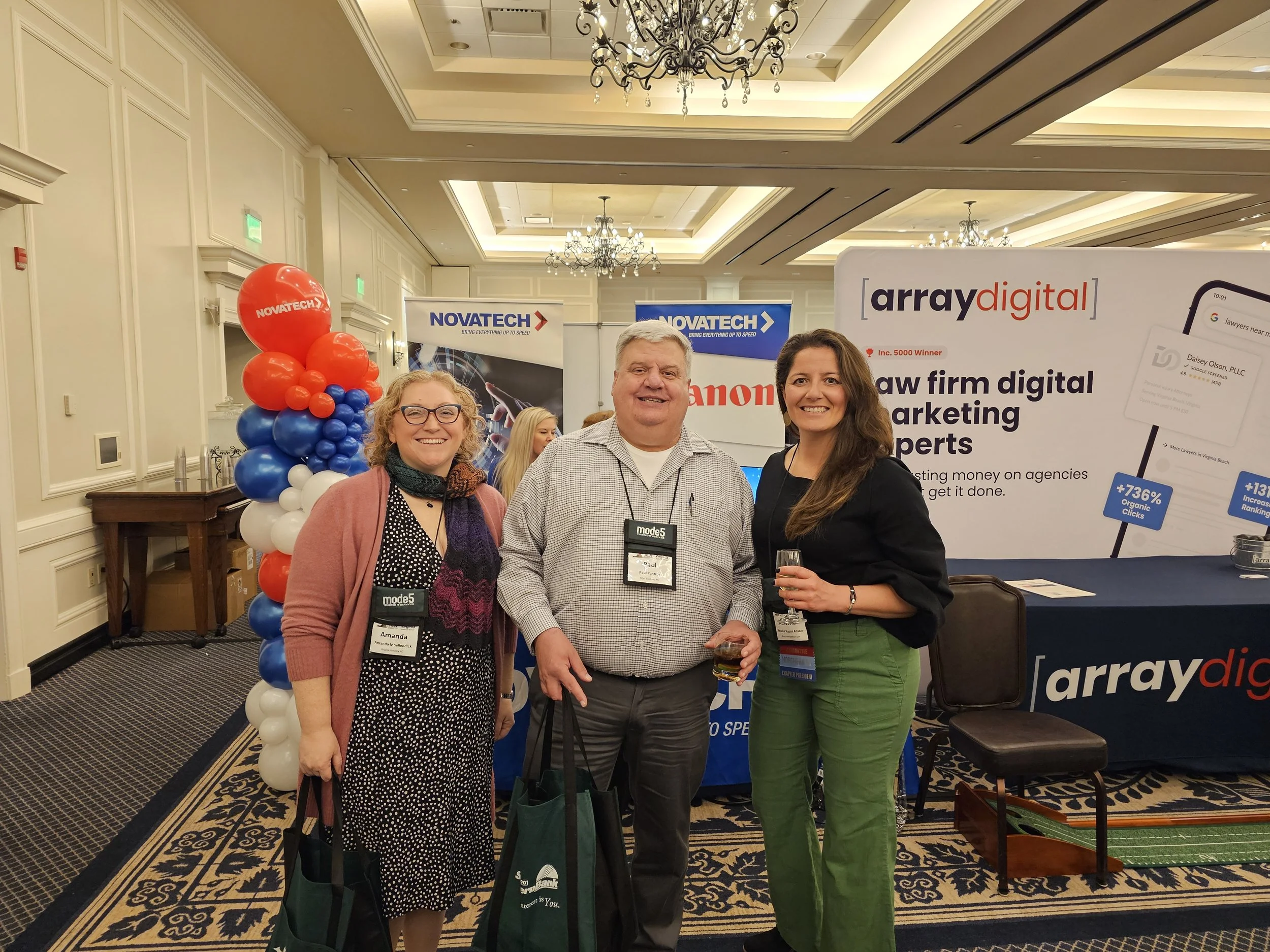 Three people standing inside a conference or expo hall, smiling at the camera. There is a woman on the left wearing glasses and a polka dot dress, a man in the middle wearing a checkered shirt, and a woman on the right with long brown hair wearing a 