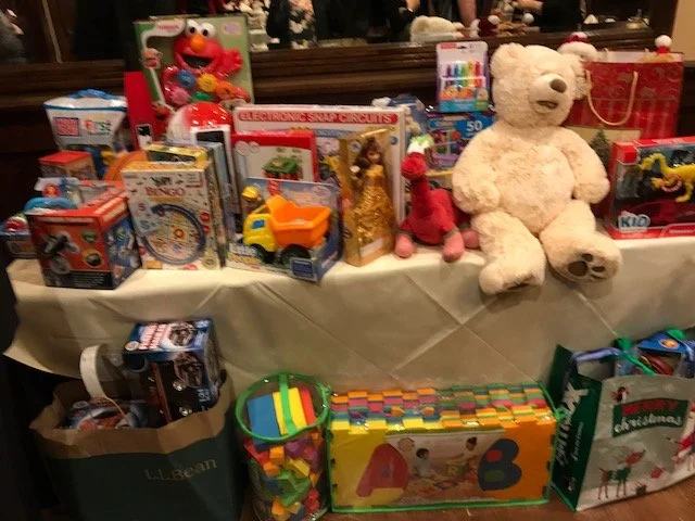 A table filled with toys including a large white teddy bear, Elmo doll, Legos, toy trucks, books, and gift bags.
