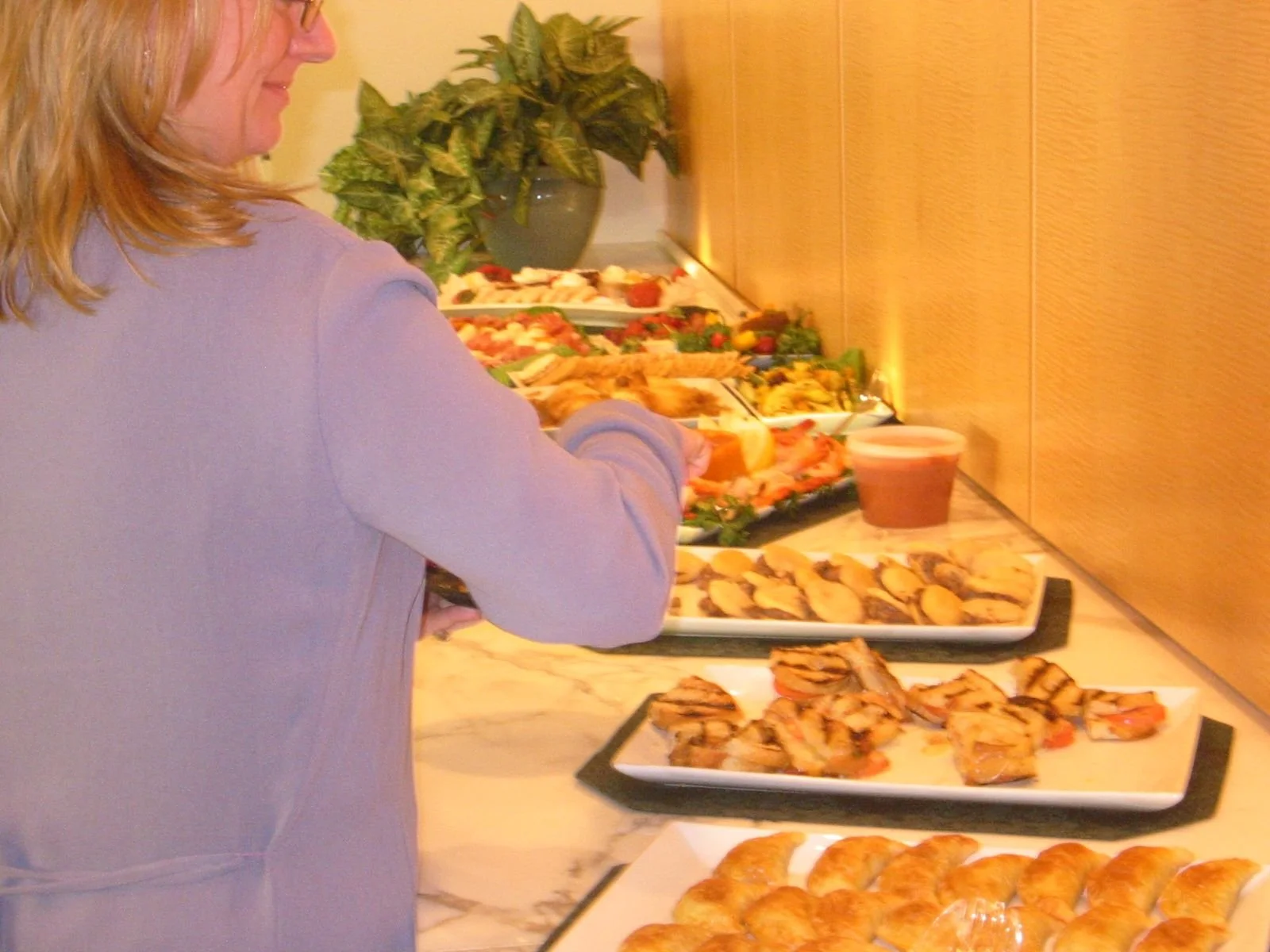 Person serving themselves food from a buffet table with various dishes including pastries, slices of meat, and salads, in an indoor setting.